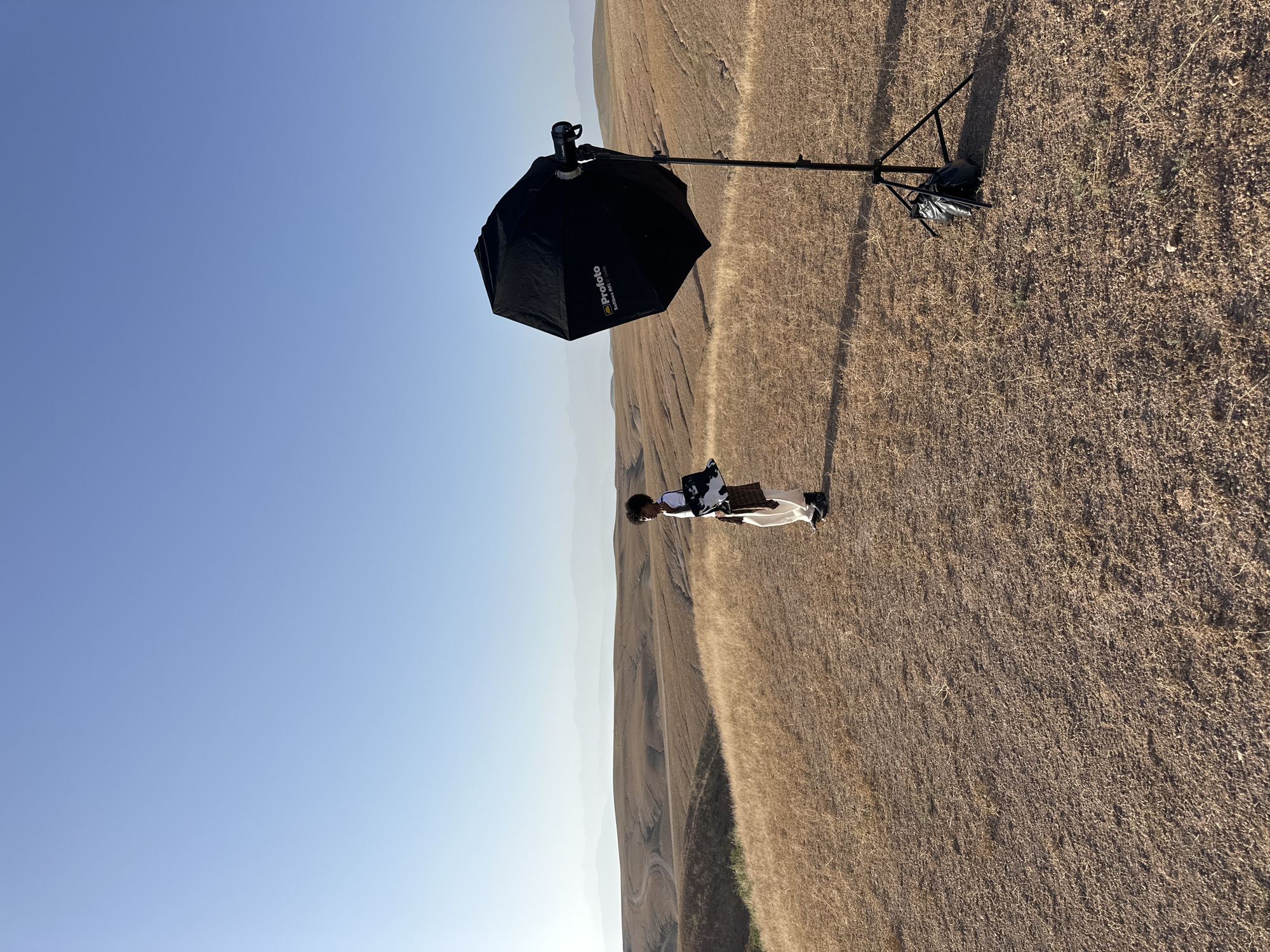 The image, taken outdoors in Sidi Badhaj, Morocco, shows a person standing in a dry, grassy field. To the left is a Profoto brand umbrella light modifier mounted on a stand. The person is wearing a white garment and appears to be holding an object. The background features rolling hills in the distance and a clear, light blue sky above. The terrain is mostly dry with sparse vegetation.