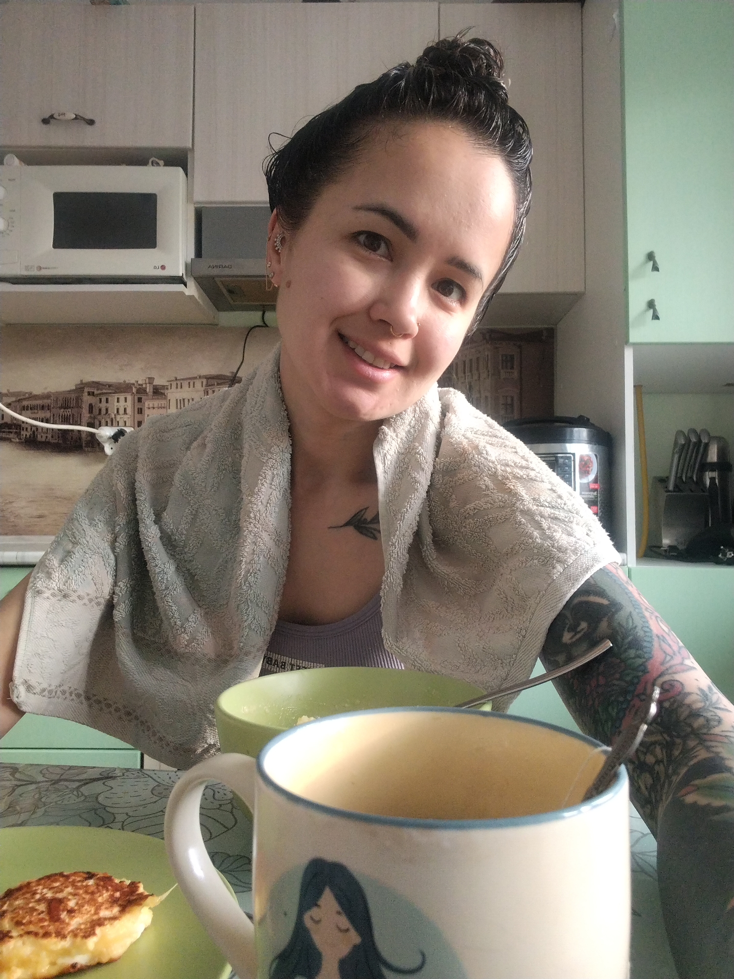 A woman smiles at the camera in a kitchen setting, with a towel draped around her neck. Her hair is pulled back in a bun, and she has visible tattoos on her chest and right arm. In the foreground, there is a breakfast scene with a slice of food on a green plate, a bowl, and a mug with a girl's face on it. A spoon rests inside the mug. The kitchen features light-colored cabinets, a microwave, and a backdrop with an image of a European city, likely Venice, Italy, not Noviy, Russia. A multi-cooker and knife block are visible in the background.