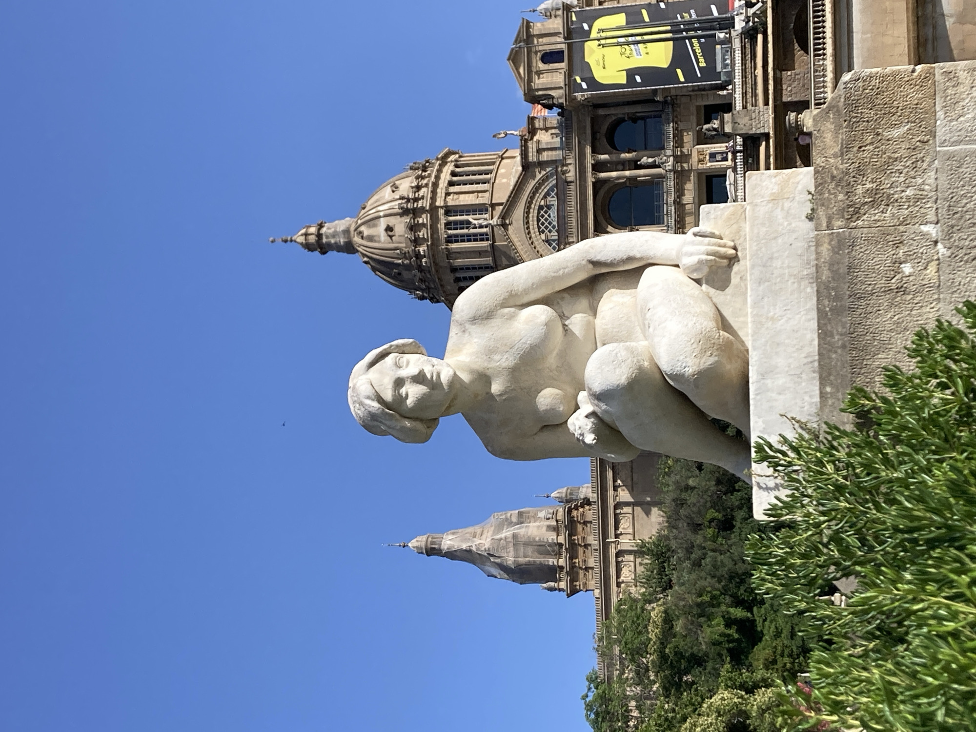The photograph captures a frontal view of a light-colored stone sculpture of a seated woman, positioned in the foreground. The sculpture is on a stone pedestal, and lush green foliage is visible in the lower portion of the frame.
In the background, the grand facade of the Palau Nacional, located on Montjuïc hill in Barcelona, Spain, is prominent. The building features a large central dome and two smaller domes, characteristic of its architectural style. A large yellow banner with black text is hanging from the building, featuring the words "Tour de France Barcelona" and other promotional text. The sky is a clear, bright blue. Two distant figures can be observed on a balcony of the Palau Nacional.