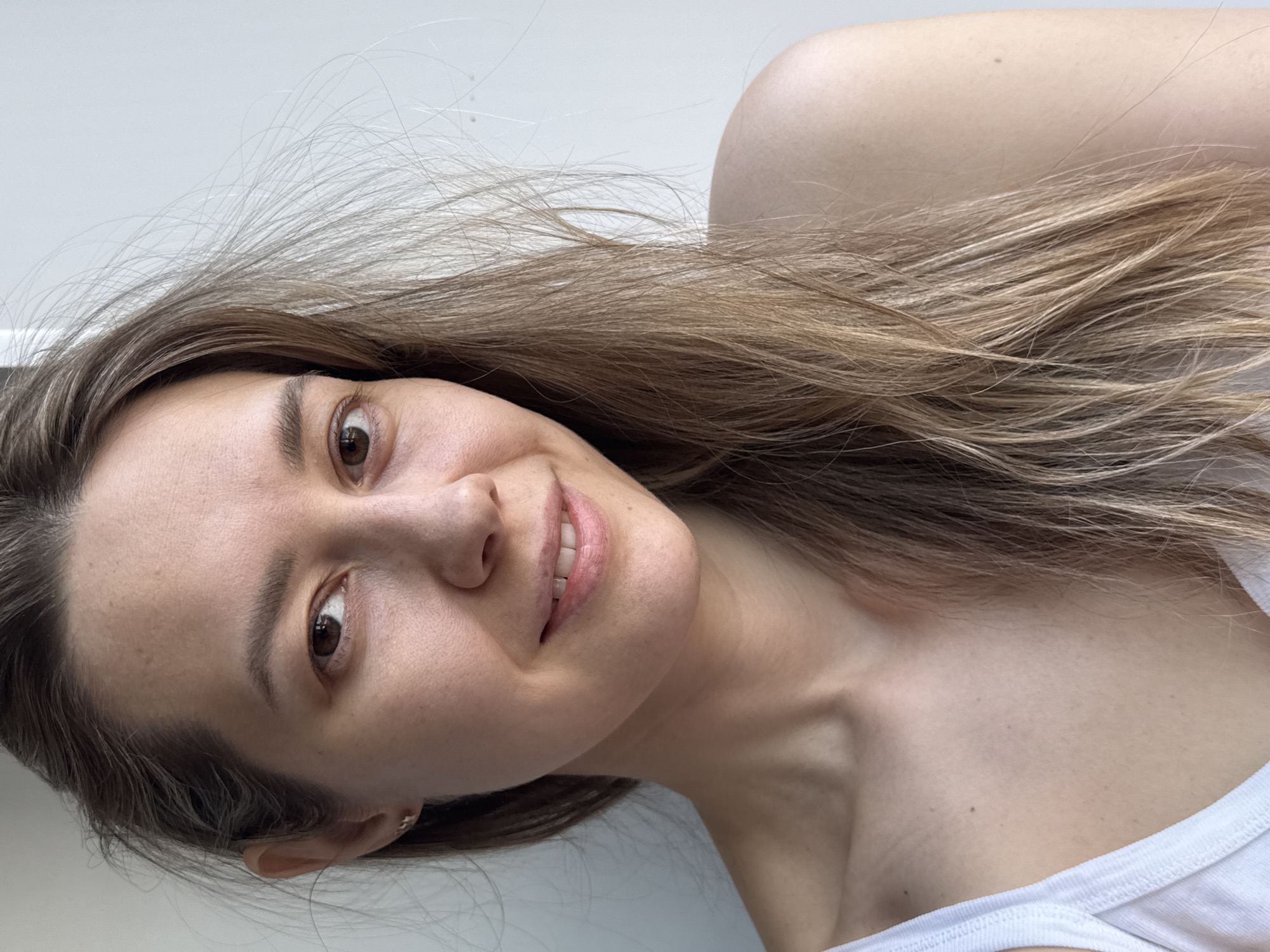 This media file presents a close-up, angled self-portrait of a young adult woman. The woman has fair skin, light brown hair with blonde highlights that appears slightly wispy, and warm brown eyes. She is looking directly at the camera with a gentle, closed-mouth smile, conveying a relaxed and friendly emotion. She is wearing a simple white ribbed tank top or camisole, visible on her right shoulder and chest. A small, subtle ear piercing is visible on her left ear.
The setting appears to be indoors, against a plain, light-colored wall that serves as a minimalist background. The lighting is soft and even, suggesting natural daylight, possibly from a nearby window, illuminating her features without harsh shadows. No specific activity is taking place other than posing for the photograph. There are no notable location cues beyond an indoor setting, and no visible text. The implied location context is Chaikovsky, Russia.