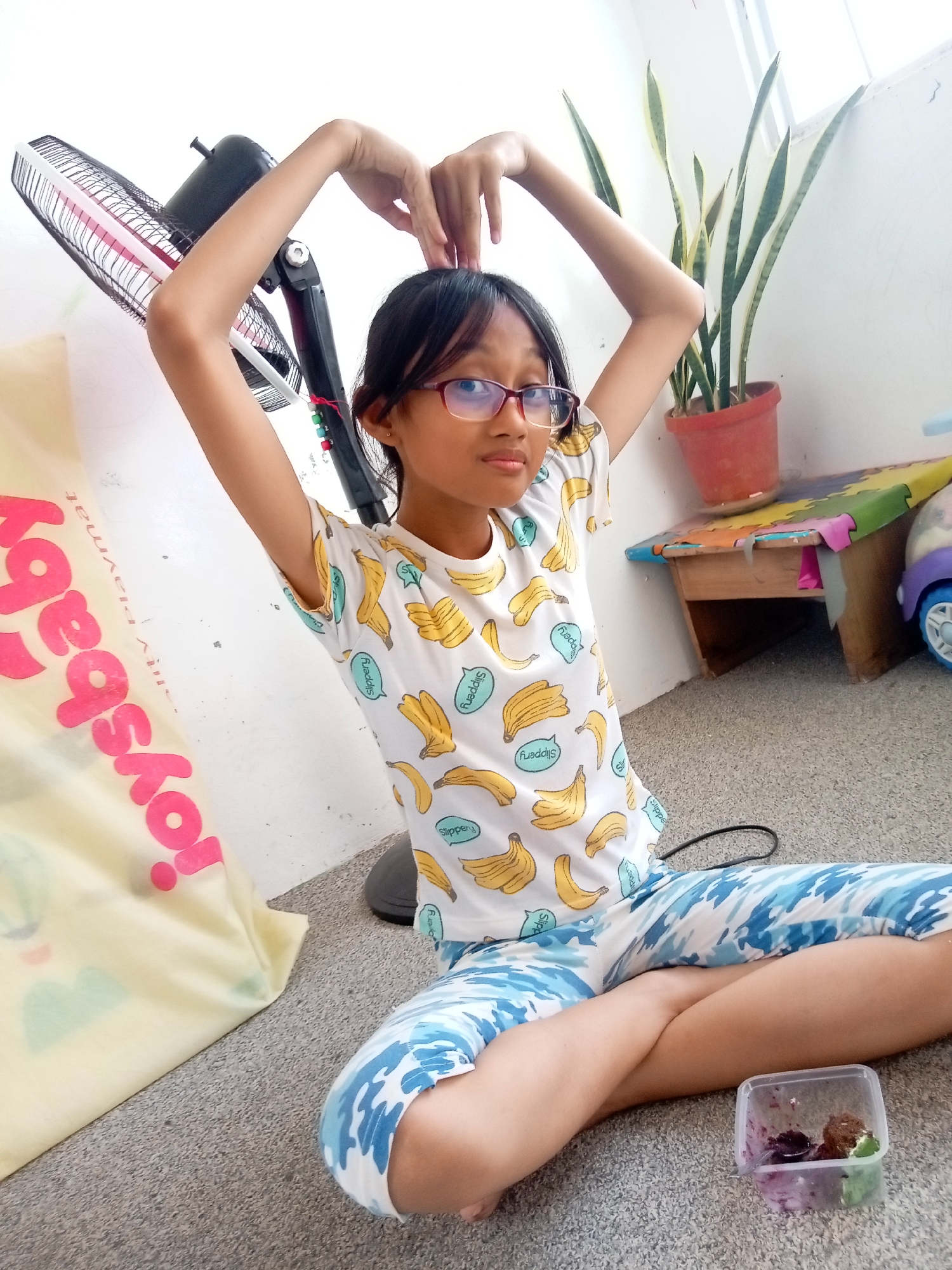 A young girl with glasses is sitting cross-legged on the floor, forming a heart shape with her hands above her head. She is wearing a white t-shirt with a banana print and blue and white patterned pants. To her left is a large yellow bag with pink lettering that reads "toys shop". In the background, there is a black fan, a snake plant in a terracotta pot, and a small wooden table with a colorful puzzle mat on top. A small plastic container with some food items is placed in front of the girl. The scene appears to be an indoor setting, possibly a home, in Batam, Indonesia.