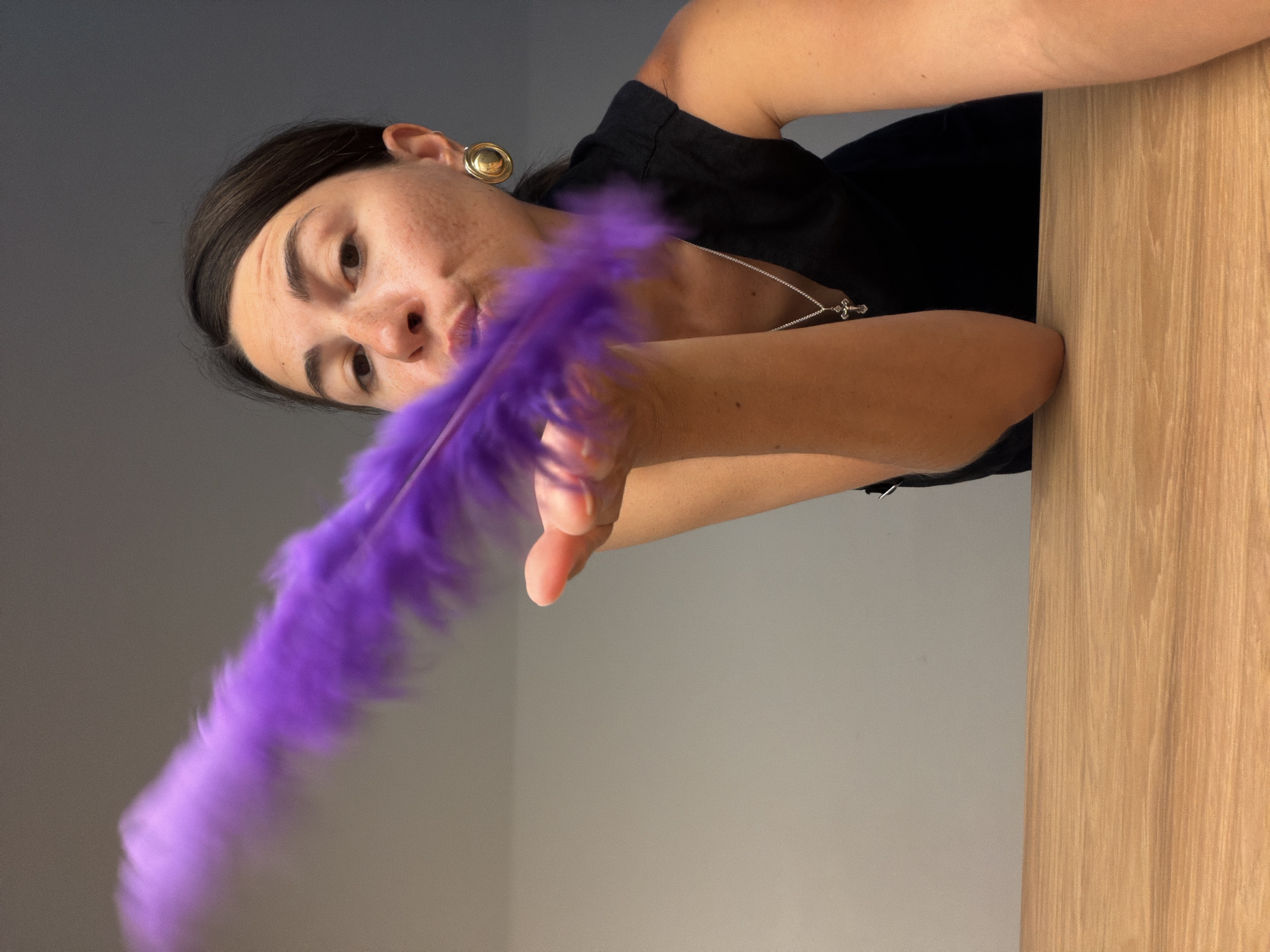 A woman leans into the frame from above, holding a vibrant purple feather. She has dark hair pulled back, a gold circular earring on her right ear, and wears a black top with a silver necklace featuring a small cross pendant. Her head is tilted, and she looks directly at the viewer with an engaging, slightly playful expression. The image is taken from a low angle, showing her upper body. The purple feather, appearing somewhat blurred, suggests motion or softness as it extends towards the lower left. The background is simple, featuring a plain grey or off-white wall on the left and a light-colored wooden surface, possibly a table or counter, on the right. The lighting is soft, indicating an indoor setting. There is no visible text in the image.