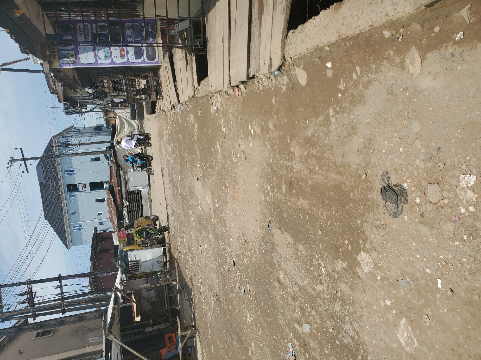 The image shows a street scene in Shangisha, Nigeria, likely during the daytime under clear skies. The ground is dirt and gravel, with scattered debris, suggesting a somewhat unkempt environment. In the background, there are several buildings, including a light blue, multi-story structure with visible windows, and other more dilapidated structures with corrugated metal roofs. Overhead, a network of electrical wires crisscrosses the sky.
On the street, two individuals are riding on a motorcycle. One person, wearing a white shirt, appears to be driving, while another person is seated behind them. Further down the street, a yellow and green motorized tricycle is parked, with what appears to be a person sitting on it, wearing a patterned skirt and green top. A sign is visible above a store or stall, displaying images of headphones and possibly mobile phones, with some text that is partially obscured. The overall impression is of a bustling, possibly commercial area in a developing urban setting.