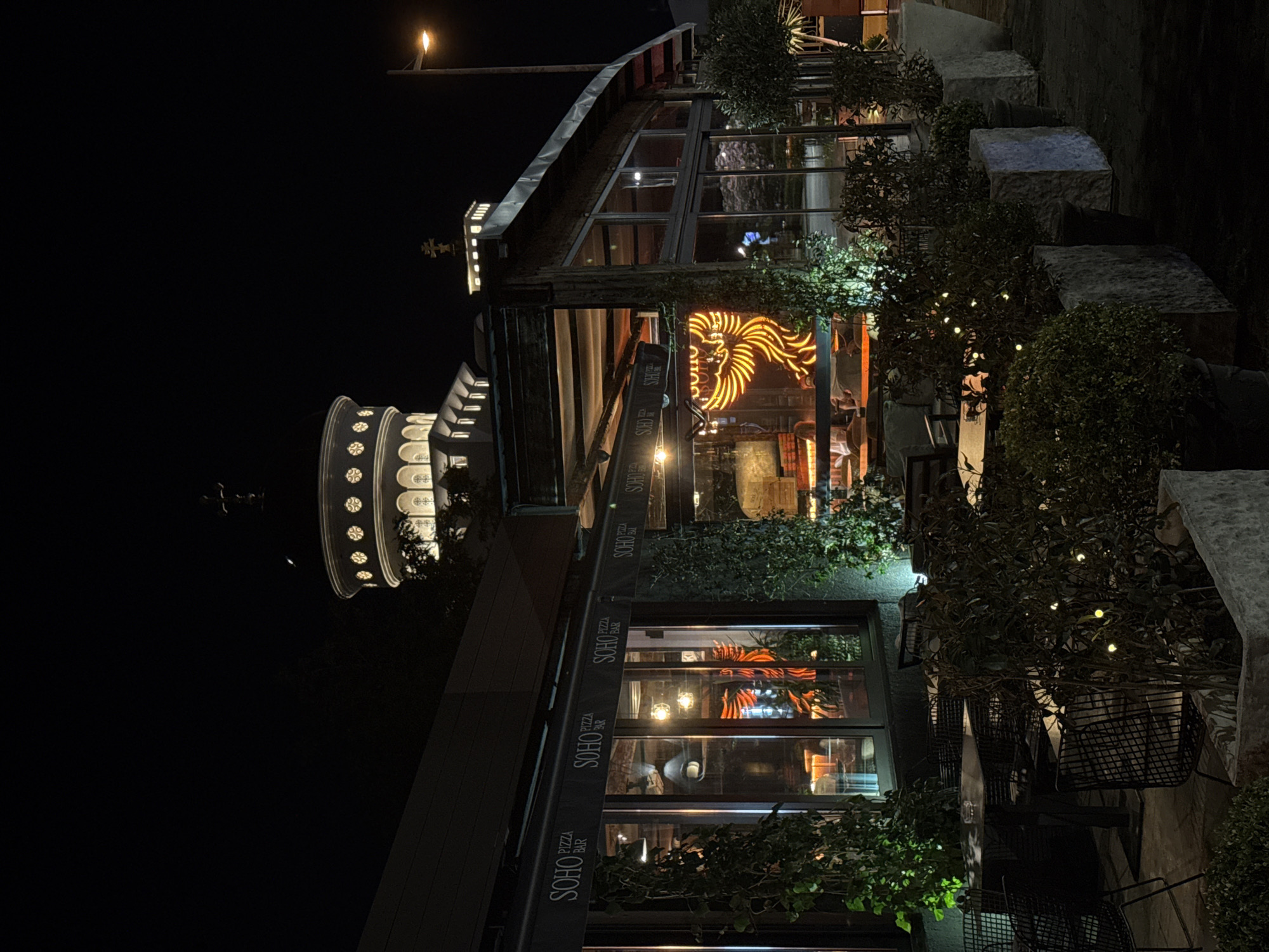 The image depicts a nighttime scene in Bar, Montenegro, showcasing the Soho Pizza Bar and the illuminated Galata Tower in the background. The Soho Pizza Bar is decorated with greenery and illuminated signs. Tables and chairs are arranged for outdoor dining. The Galata Tower is visible in the distance, illuminated against the night sky.