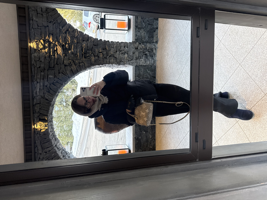 A person is reflected in a large framed mirror/glass door, taking a selfie with a white iPhone held in their right hand. The individual is wearing a black puffer jacket, black pants, black gloves, dark-rimmed glasses, and a light beige scarf draped over their head and neck. Dark blue suede ankle boots are visible on the light-tiled floor. A small Louis Vuitton Monogram handbag, featuring a gold chain and shoulder strap, is carried in the left hand. The background reflected in the mirror depicts a building interior with dark grey stone archways illuminated by recessed spotlights. Through the archways, an exterior scene in Courchevel, France, is visible, showing snow-covered ground, evergreen trees, a paved road, and a white vehicle partially obscured on the right. Two tall, dark-framed lanterns with orange internal illumination flank the archway, and a large, dark brown amphora-shaped pot stands to the left.