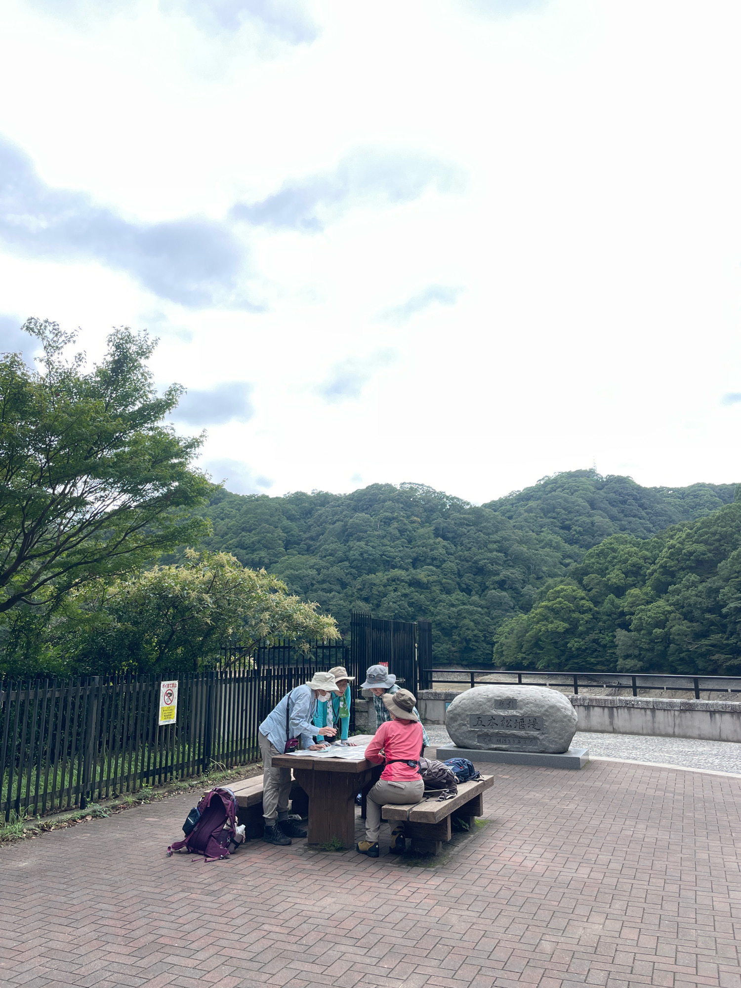 Four individuals, dressed in casual outdoor attire and hats, are gathered around a wooden table and bench. Three of them are looking down at a map spread across the table. One person is seated on the bench, facing away from the camera, while the other two are standing. A fourth person is partially visible, seated on the bench to the right. A purple backpack and another bag are on the ground near the bench.
The scene is set outdoors on a paved area with a brick pattern. To the left, a black metal fence separates the paved area from a grassy embankment with trees. In the background, a lush, tree-covered mountain range dominates the horizon under a cloudy sky. A large, inscribed stone monument stands to the right of the group. A body of water and a concrete barrier are visible beyond the monument, suggesting a dam or reservoir. The location is identified as Kobe, Japan, specifically the Kasagayama Dam (笠置山ダム).