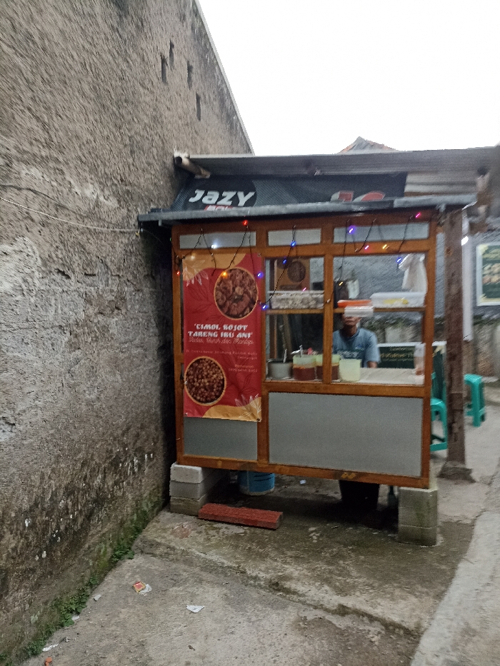 The image shows a small food stall in Jatinangor, Indonesia, located at the coordinates -6.934 latitude and 107.775 longitude. The stall is made of wood and has a gray metal roof. There is a sign above the stall that reads "Jazy". The stall has a window where a person appears to be working. There is a sign on the front of the stall with the words "CIGOL SOJOT TARENG IRU ANY" and a picture of food. There are strings of lights hanging above the stall. There are several concrete blocks beneath the stall and a brick path in front of it. The stall is located in a narrow alleyway with a gray concrete wall on the left side of the image. The alleyway is paved with a light gray concrete surface. There is some debris and green vegetation on the ground. The ground is wet. The sky is overcast and there is a dark roof in the background. The time of day is likely afternoon or evening.