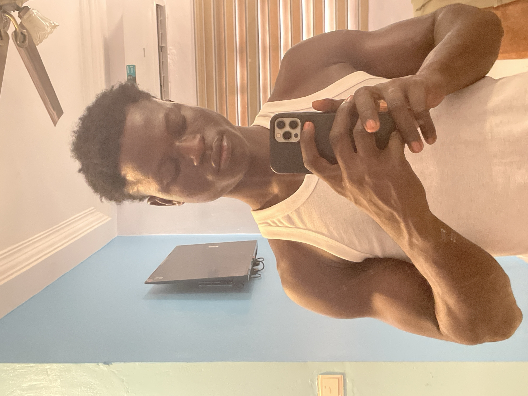 A dark-skinned man in a white tank top is taking a selfie with a black smartphone. He is indoors, possibly in a room in Badagry, Nigeria. There is a laptop on a light blue surface and a ceiling fan in the background. There are white or beige vertical blinds in the background, next to a white wall. A light switch is visible near the bottom of the frame. The walls are painted light blue and off-white.