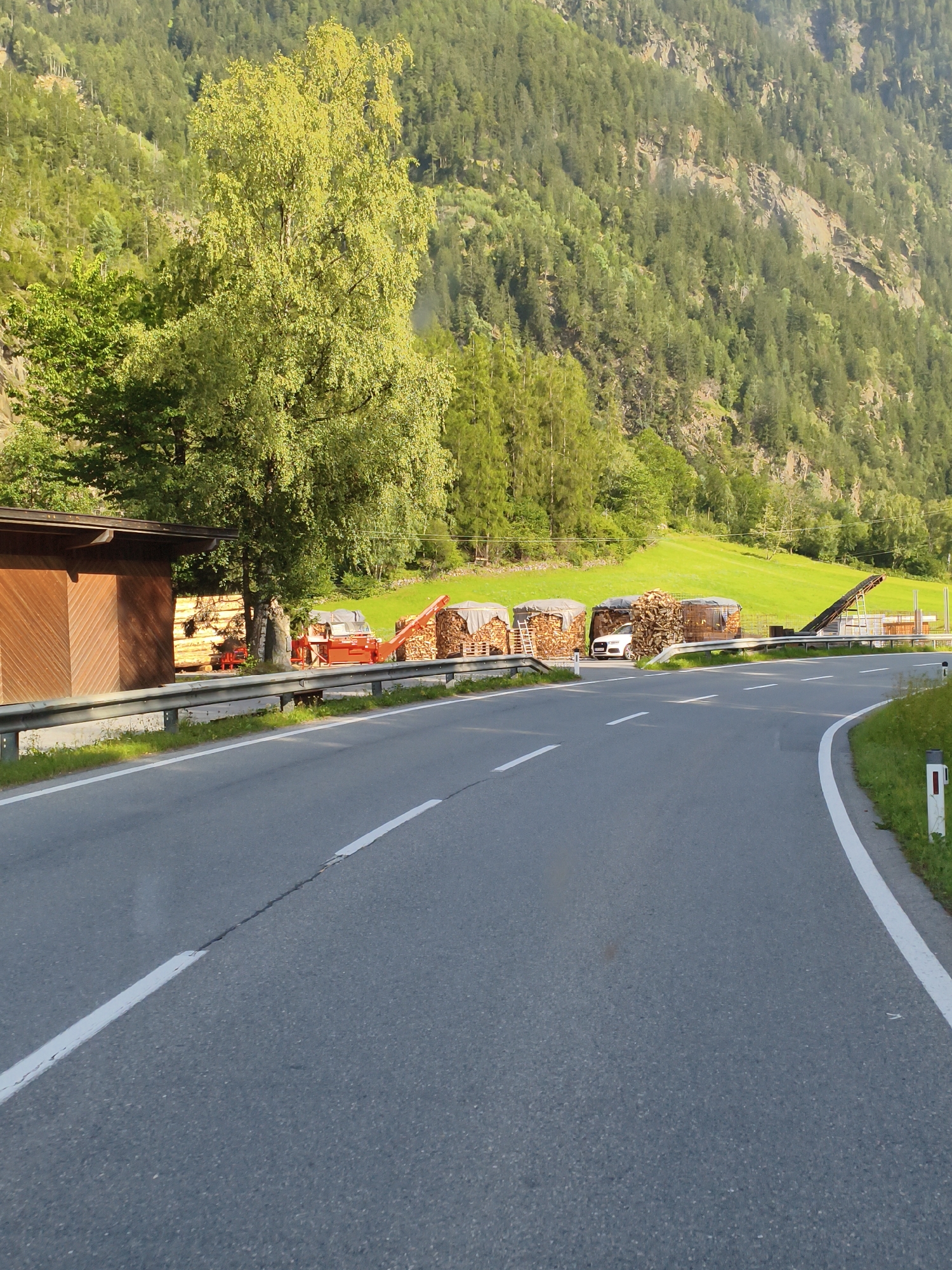The image depicts a road curving through a mountainous landscape in Tumpen, Austria. The foreground is dominated by the grey asphalt road with white lane markings. To the left, a wooden building with a pitched roof is visible, adjacent to a pile of logs. Further down the road, to the right of the building, are several stacks of firewood covered with tarps, some of which are on pallets. A white car is parked near one of these stacks. In the background, a steep, tree-covered mountainside rises, with a bright green meadow visible partway up. A metal guardrail runs along the side of the road. The lighting suggests it is daytime, with sunlight illuminating the scene. There are no people visible in the image.