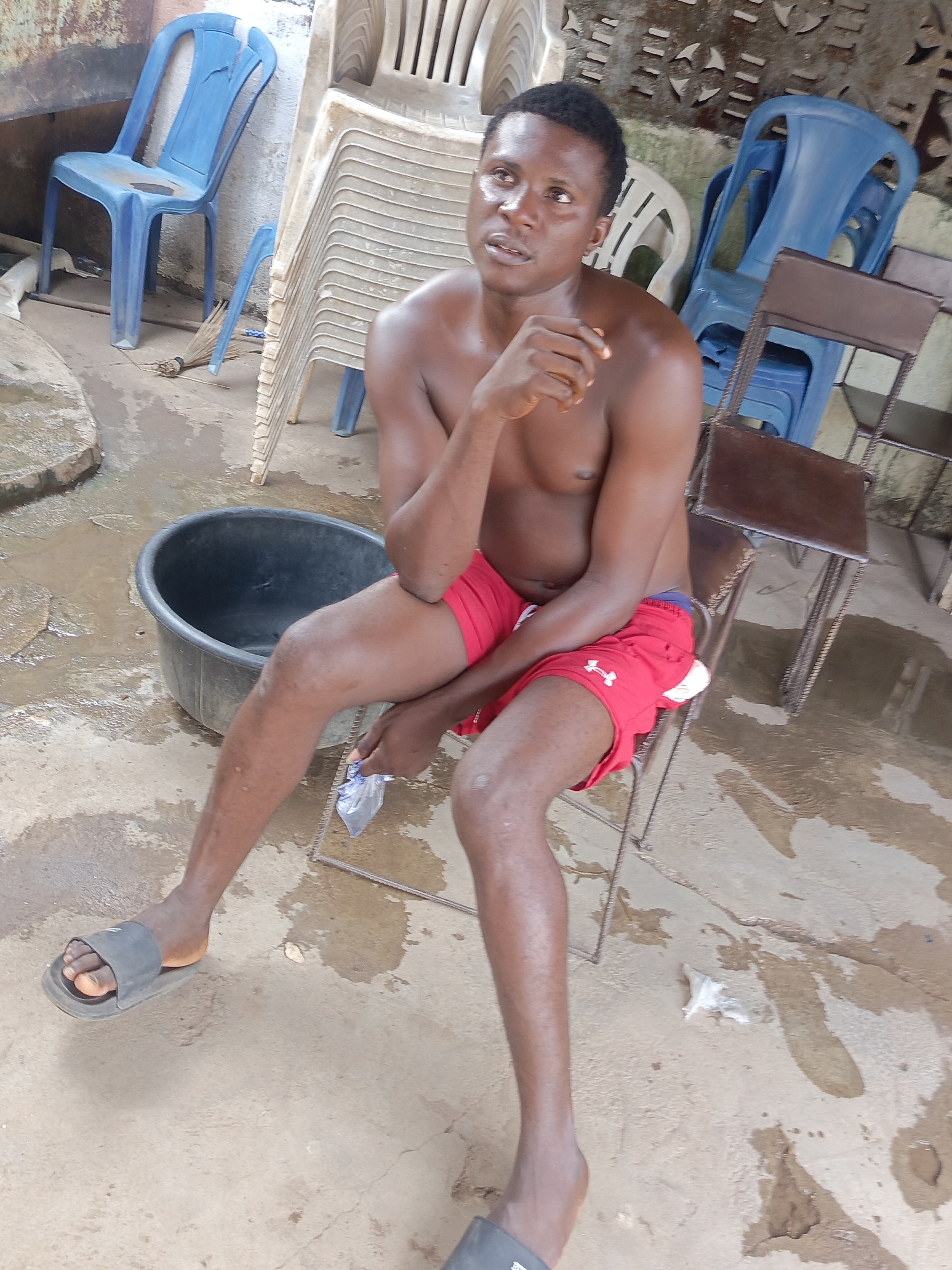 A man sits shirtless in a chair outdoors in Unguwar Zaria, Nigeria. He wears red shorts and dark-colored slides. He is dark-skinned, with his left hand raised near his face. He holds a small plastic bag in his right hand. A black tub sits to his left. Several plastic chairs are stacked behind him, with additional chairs of different styles surrounding him. The ground appears wet.
