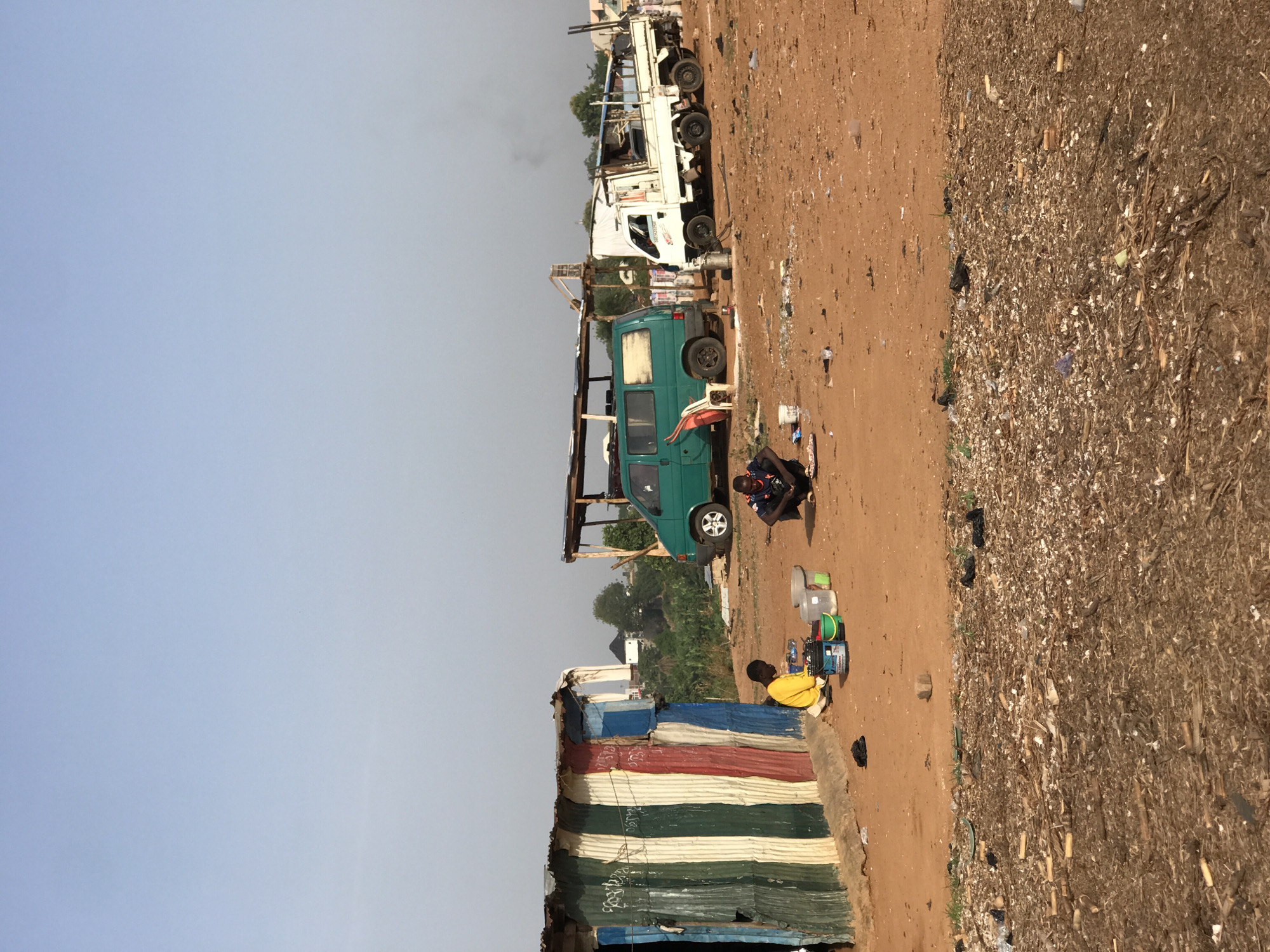 This outdoor scene, set in Abuja, Nigeria, depicts an active, semi-urban or informal environment under a hazy, pale blue-grey sky. The ground is a reddish-brown dirt path on the left, transitioning to a much darker, textured area covered in dry organic matter, possibly agricultural waste or general refuse, on the right.
In the foreground on the left, a makeshift structure, likely a dwelling or stall, is constructed from horizontally arranged corrugated metal sheets painted in stripes of blue, red, green, white, and yellow. Visible on these sheets are handwritten numbers and text, including "2023" repeated several times.
Further back and to the right, a teal-green van, possibly a Volkswagen Transporter, is parked under a simple wooden and corrugated iron canopy. A white plastic chair sits next to its front wheel. Behind the van, a larger white truck, appearing to be a flatbed, is visible, with a faint plume of smoke rising into the sky.
Two individuals are seated or crouching on the dirt ground in the mid-ground, towards the right. One person, wearing a yellow top, is facing away from the viewer and interacting with several plastic containers of varying sizes and colors (white, green, blue, clear). Another person, in a dark top with blue patterns, is seated nearby, also facing the containers. Their posture and the containers suggest they are engaged in a task such as sorting, cleaning, or preparing goods. The scene occurs during daytime, under clear but hazy weather conditions, and reflects a functional, informal setting.