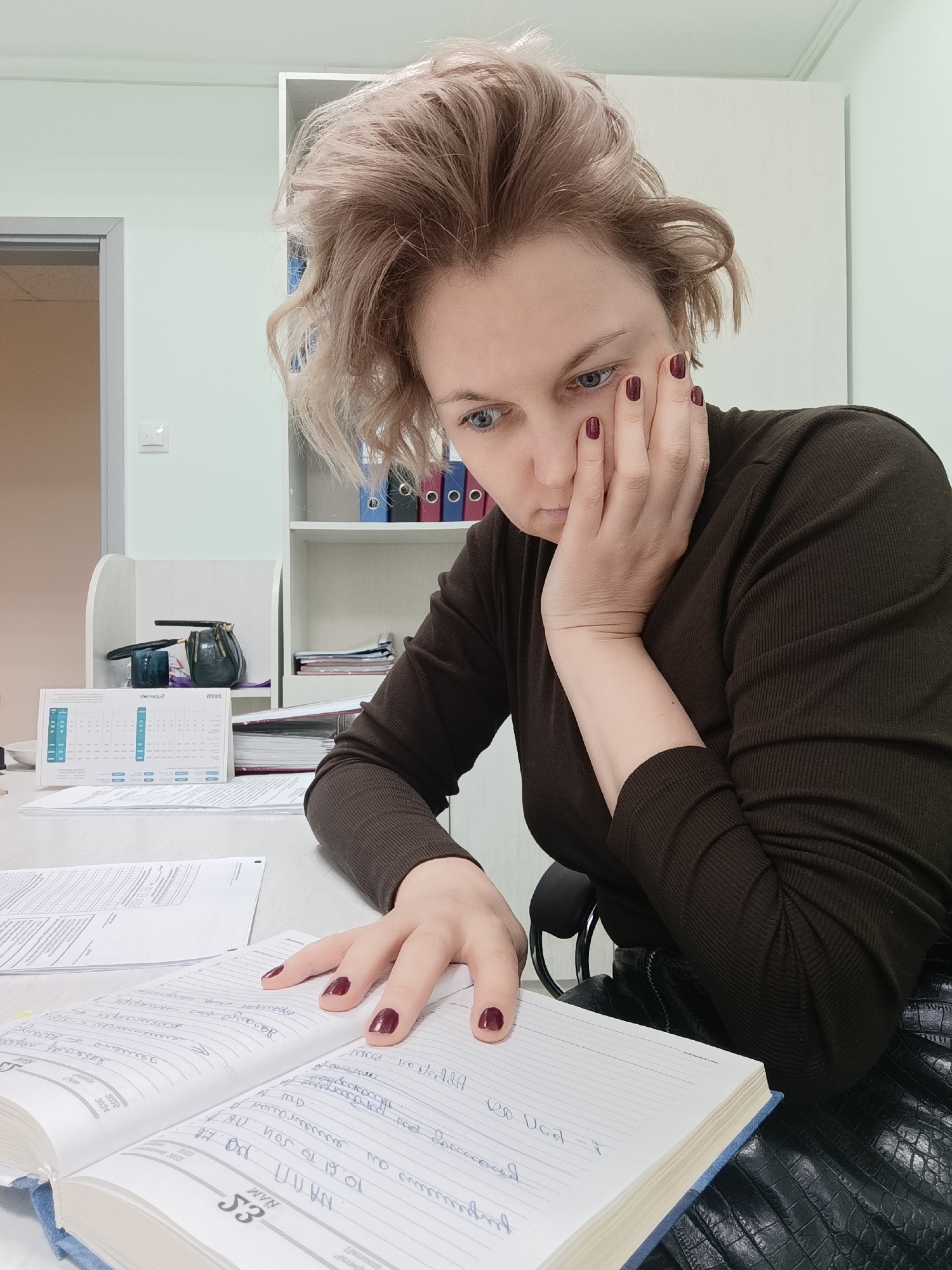 A woman with blonde hair and burgundy nail polish sits at a desk, looking down at an open notebook. She rests her chin on her hand in a pensive pose. The notebook is filled with handwritten notes in a script. The desk also contains loose papers, a calendar, and a small, dark-colored purse. A white bookshelf with files is in the background. A door is visible in the top left corner of the frame. The image was taken in Марковский, Russia.