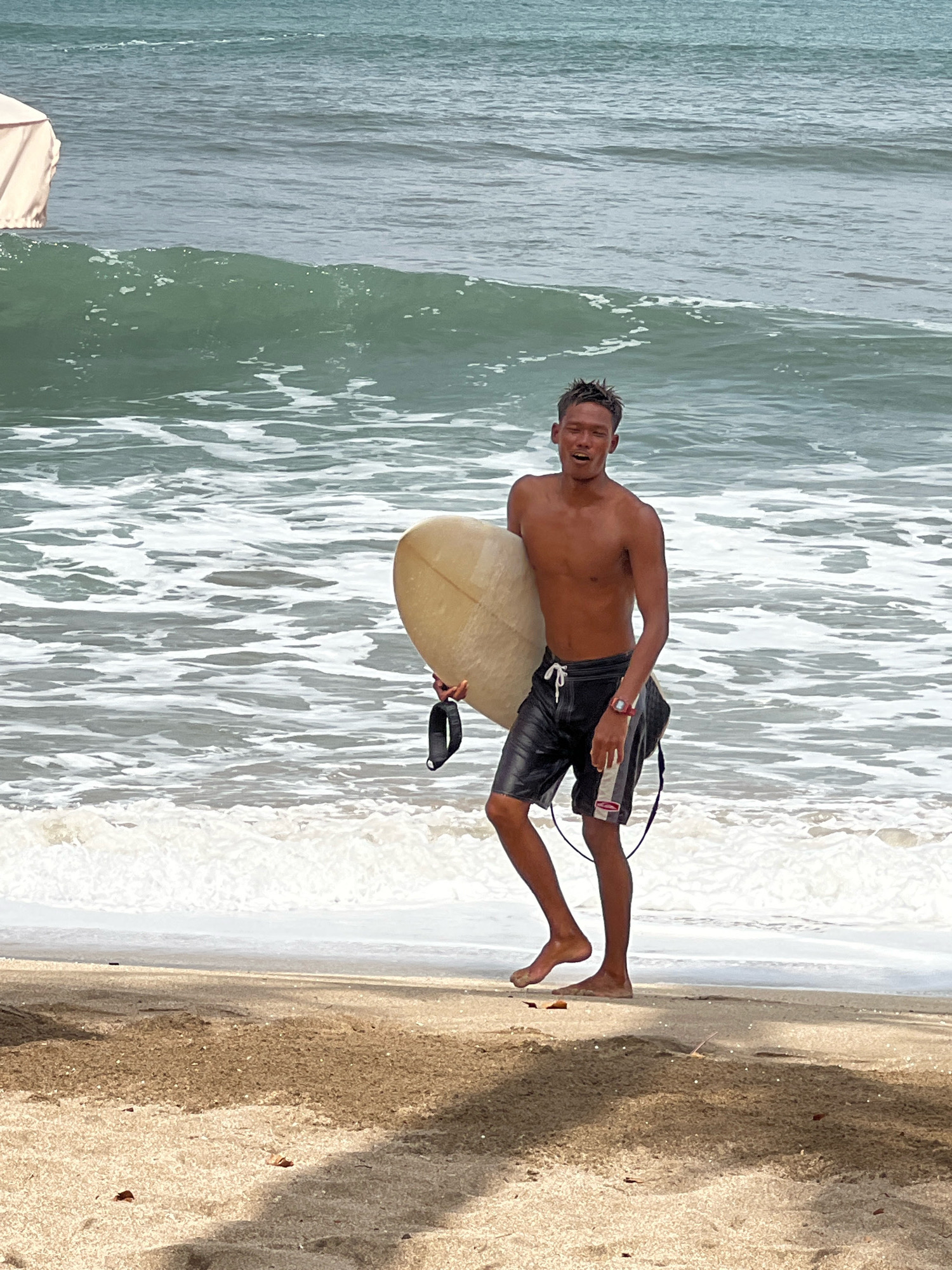 A young, shirtless man is walking out of the ocean on a sandy beach in Kuta, Indonesia, carrying a light-colored surfboard. He is wearing black swim trunks with white and grey stripes. A black mask hangs from his right side. The ocean behind him has waves with white foam. The sky is overcast.