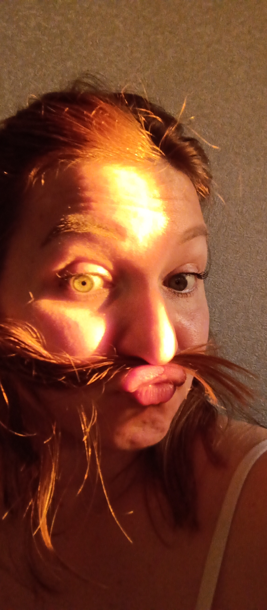 A close-up shot depicts a woman indoors in Unknown, Unknown. She has reddish-brown hair with strands arranged to resemble a mustache. The woman has one eye visible on the right side of the frame, with the left eye being hidden by the hair. She's pulling a face with pursed lips, and her hair is used to create the illusion of a mustache above her mouth. A strong light source illuminates her face, causing an uneven distribution of light and shadow. The background is a plain wall, and a portion of her clothing is visible.