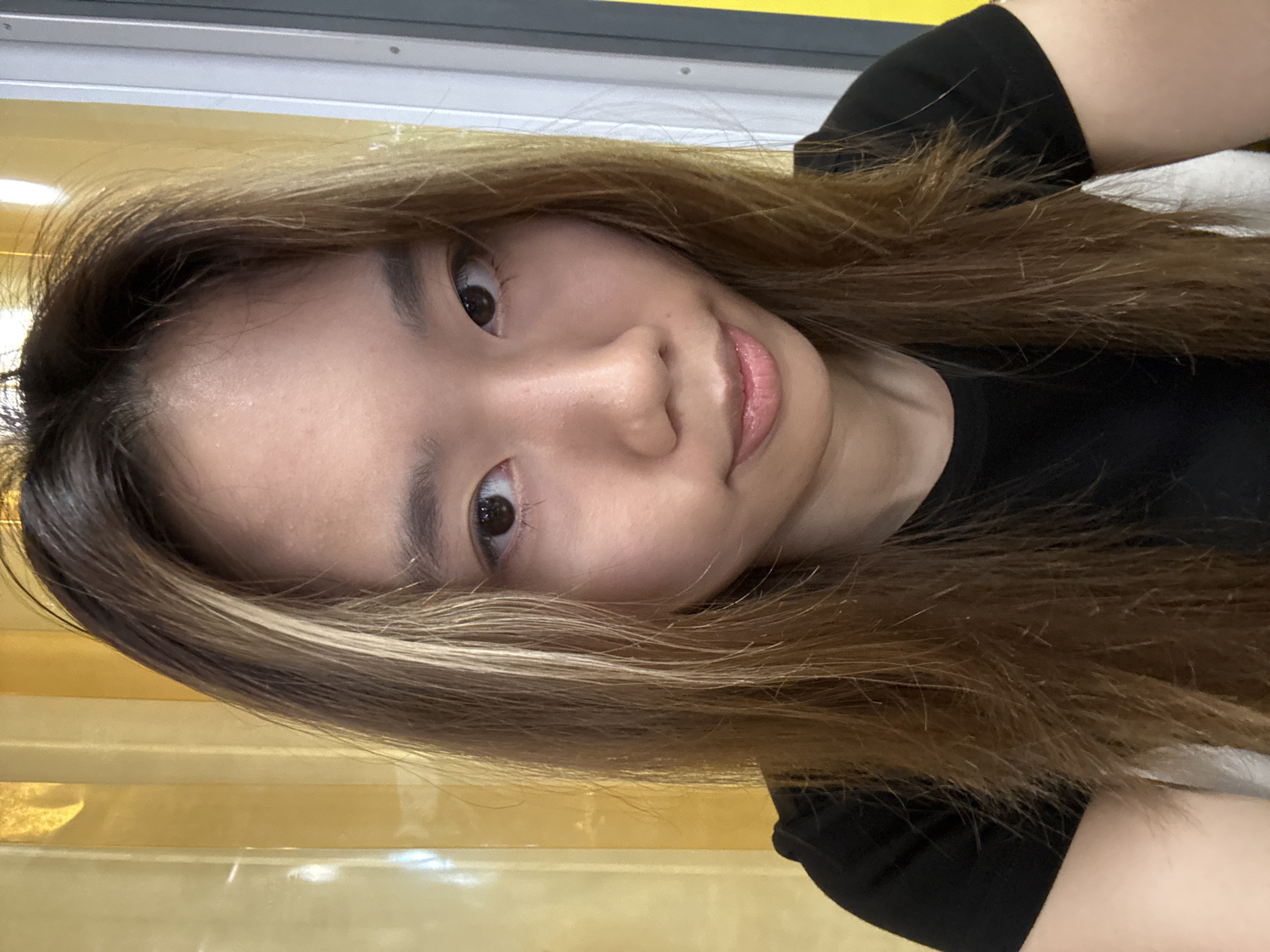 A young woman with shoulder-length brown hair with blonde highlights is posing for a selfie. She is wearing a black t-shirt. The background is a blurred combination of yellow and grey.