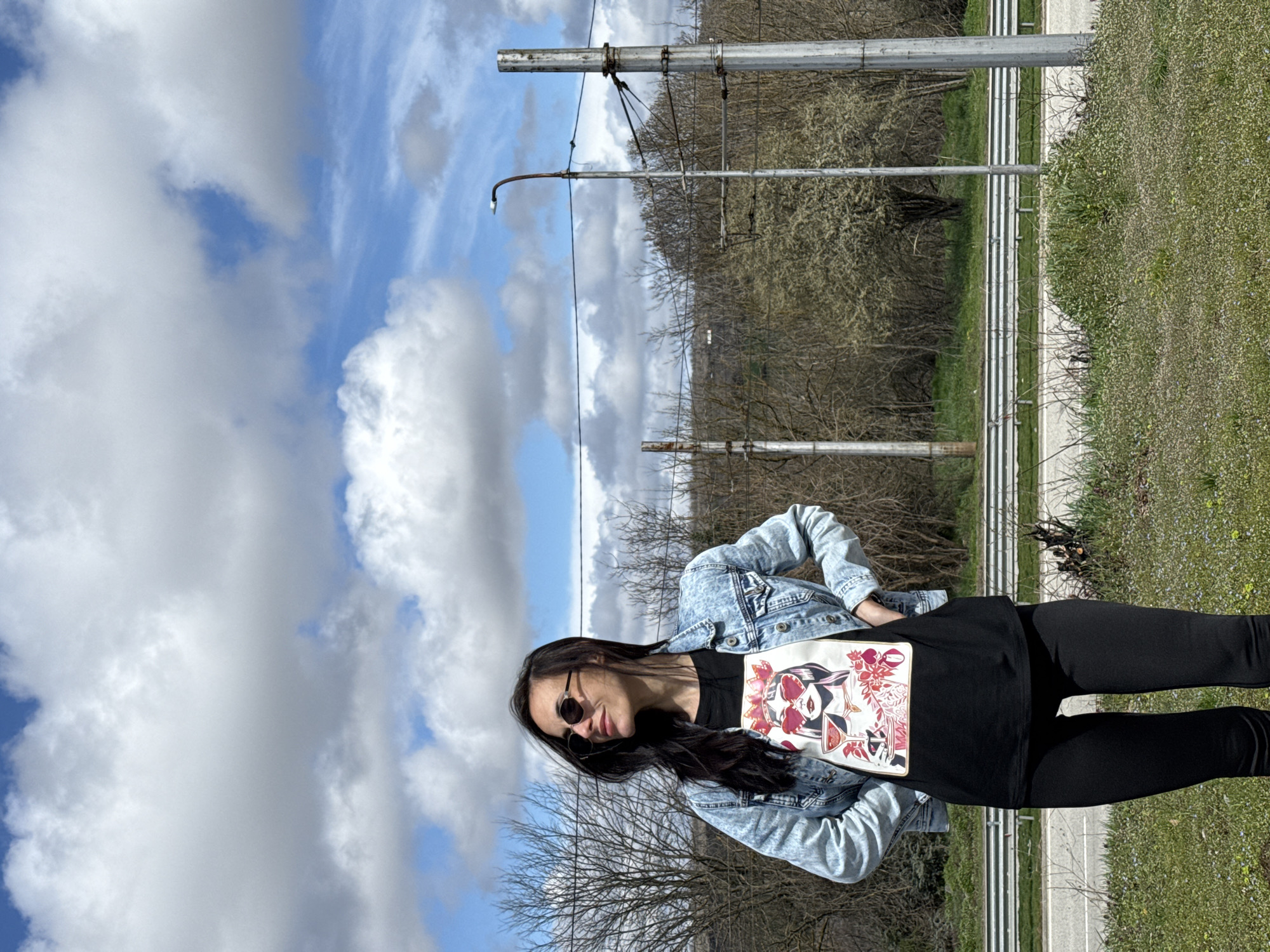 A single female subject stands outdoors in Pleven, Bulgaria, under a blue sky with scattered white clouds. She has long dark hair and wears dark, oval-frame sunglasses. Her attire consists of a light-wash denim jacket, a black t-shirt featuring a stylized graphic of a woman with heart-shaped glasses holding a cocktail, and black leggings. Her hands are placed on her hips, and her head is turned to the viewer's right. The immediate foreground displays green grass with small flora. Behind the subject, a metal road barrier and an asphalt road are visible. The background features several bare deciduous trees and shrubs, along with multiple galvanized steel utility poles supporting overhead wires. One pole is equipped with a curved street light arm and bulb. Distant terrain is visible beyond the vegetation.
