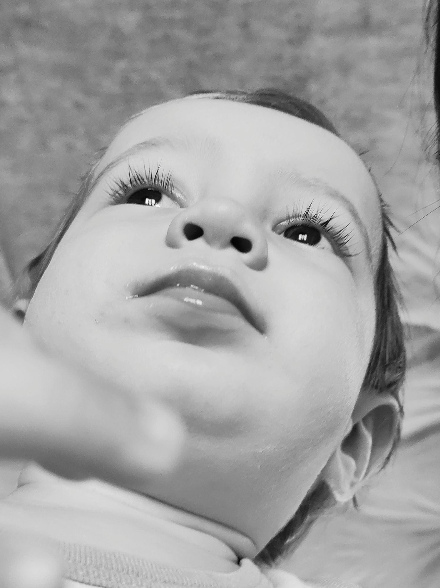 A close-up, black and white photograph depicts a baby lying on its back. The baby is fair-skinned and has long eyelashes. The baby's lips are slightly parted. The baby's gaze is directed upwards. A blurred object, potentially a hand or object, is in the foreground of the image. The background appears to be a neutral tone, possibly fabric or bedding. The location is unknown and not mentioned within the image and therefore cannot be Фоки, Russia.