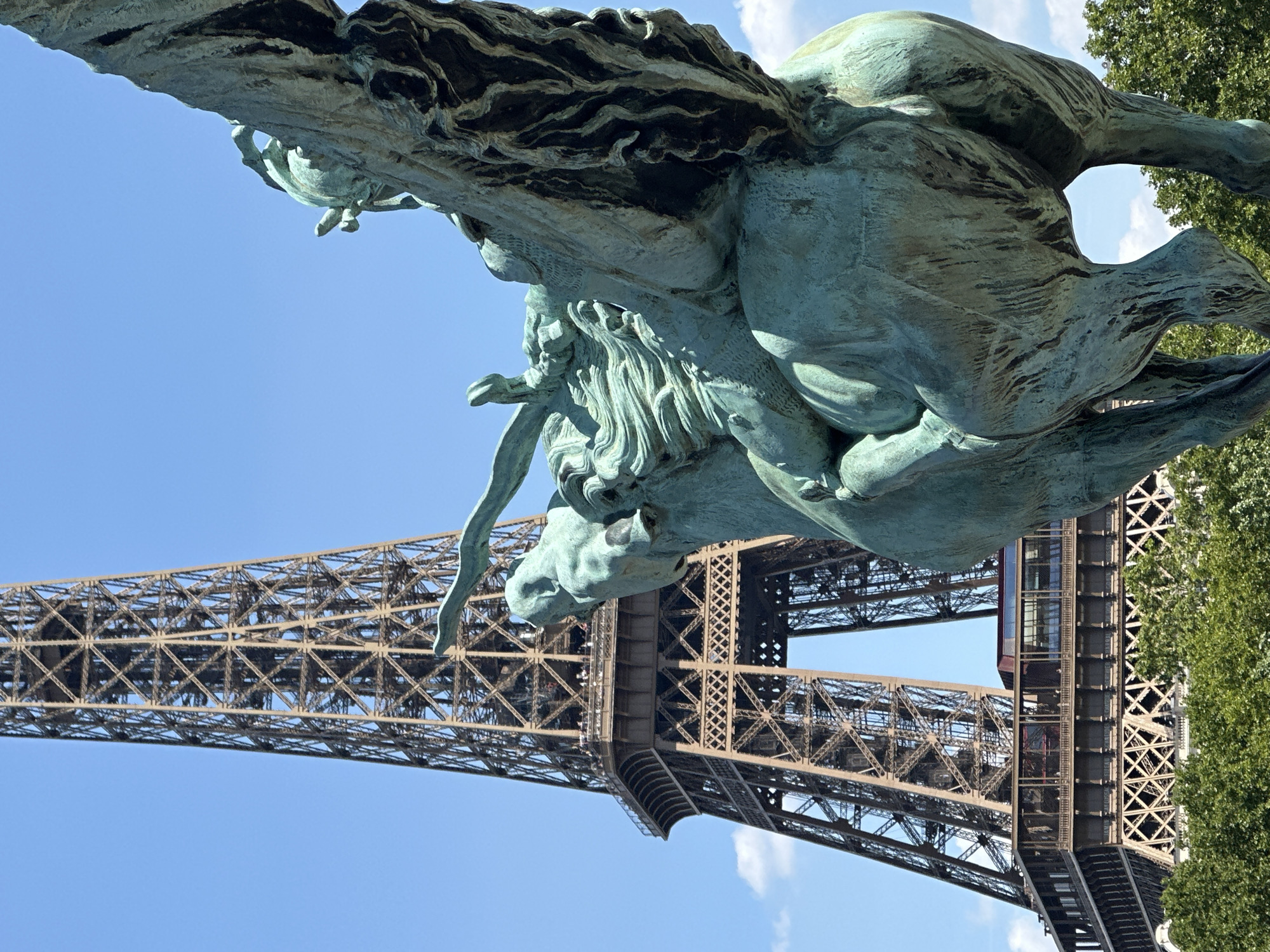 The image shows a close-up of a bronze statue of a winged horse with a rider, partially obscuring the Eiffel Tower in the background. The statue, weathered to a green patina, is positioned in the foreground, with its intricate details of musculature and mane visible. The Eiffel Tower, a landmark of Paris, France, is seen in the mid-ground, its lattice structure clearly defined against a bright blue sky with sparse white clouds. The lower portion of the tower is surrounded by lush green trees. The statue's perspective suggests it is part of a larger monument, possibly located in a park or public square near the Eiffel Tower. No people are clearly discernible on the statue itself.
