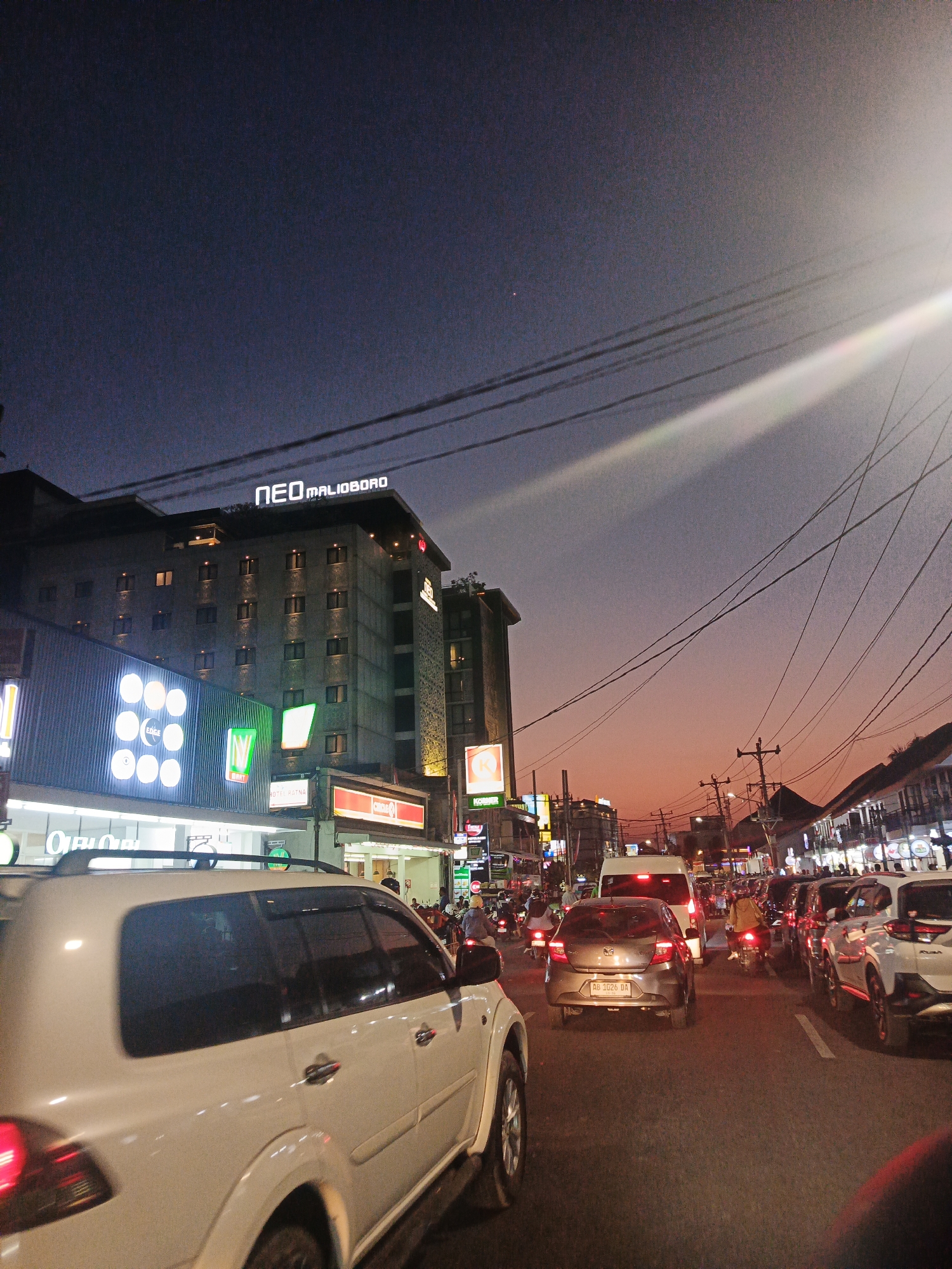 This is a photograph of a street scene in Yogyakarta, Indonesia, at dusk. The sky is a gradient of deep blue to orange, with power lines crisscrossing the frame. In the background, a large, multi-story building with the illuminated sign "NEO MALIOBORO" is visible. To the left of the building, there are several illuminated signs, including one with circular lights labeled "EDGE." The street is congested with traffic, including cars and motorcycles. In the foreground, the side of a white SUV is prominent. The overall impression is of a busy urban environment at the transition from day to night, likely a commercial or hotel district given the signage.