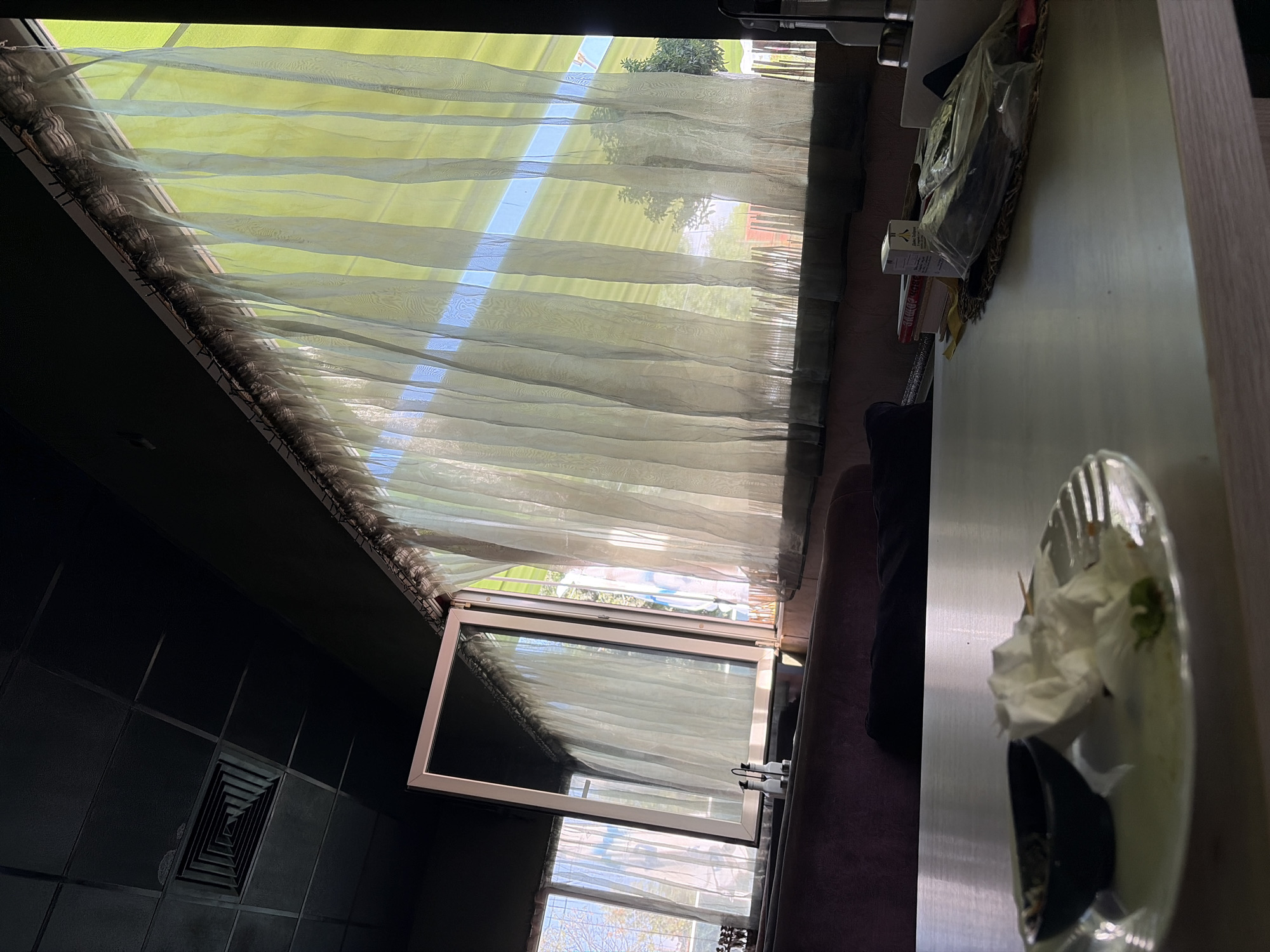 The image shows a table in what appears to be a restaurant or cafe. The table has remnants of a meal, including a plate with used napkins and a small bowl. On the table, there are also some books, a box, and plastic-wrapped items. The background features a large window covered by sheer curtains, allowing sunlight to filter through. Outside the window, trees and fencing are visible. The ceiling is dark and features a square air vent. The overall impression is that of an indoor dining space with natural light coming from a window.