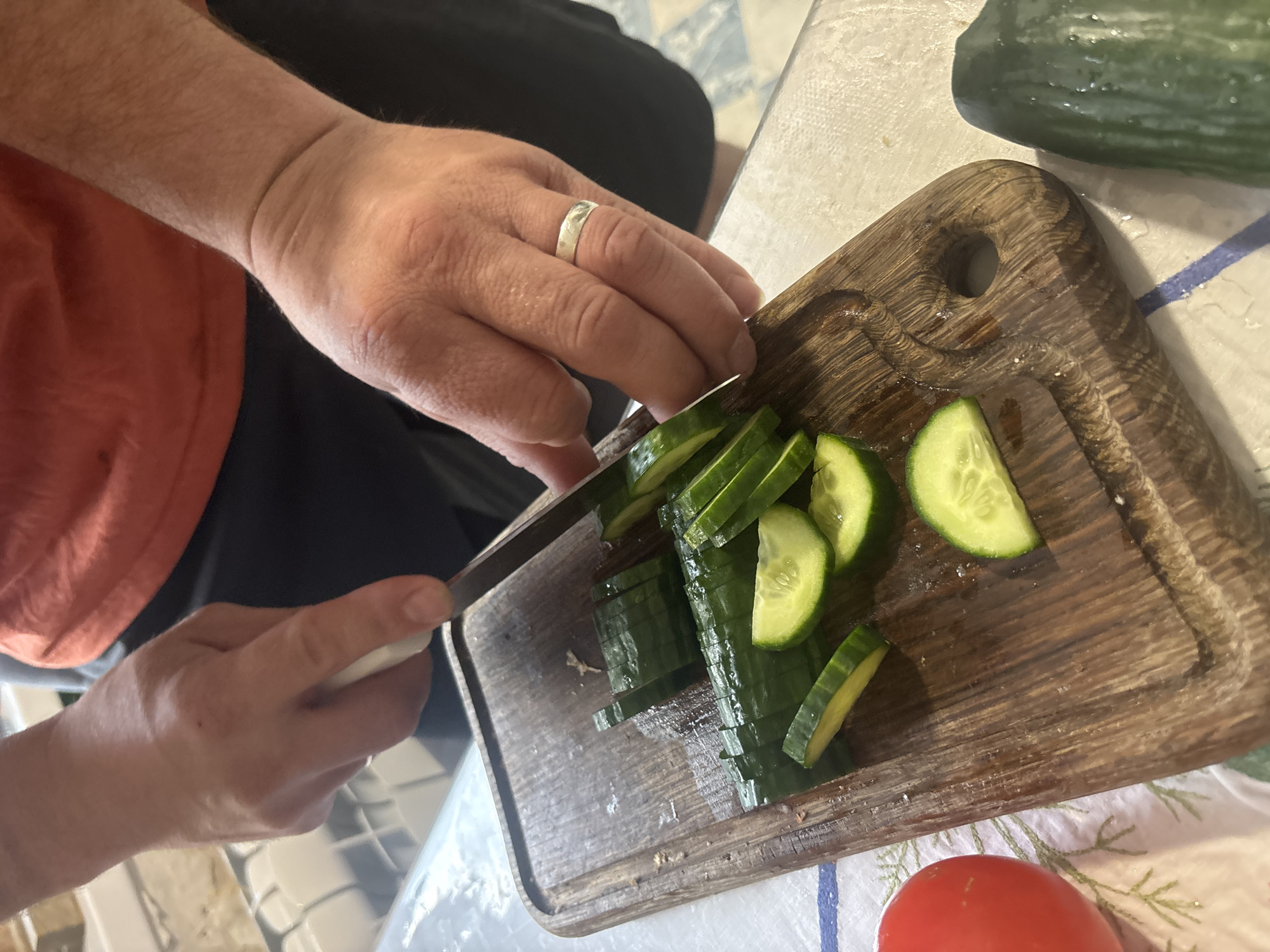 The image presents a close-up, high-angle view of a person's hands actively engaged in slicing a cucumber. The central subject is the hands of an individual, likely male given the size and the presence of a silver band-style ring on the ring finger of the left hand. The person is wearing an orange or terracotta-colored short-sleeved shirt and dark shorts or trousers.
The left hand is holding a kitchen knife with a white handle, positioned over a partially sliced cucumber. The right hand, though partially obscured, appears to be steadying the cucumber during the cutting process. Several neatly cut, thin slices of cucumber are visible on a sturdy, dark brown wooden cutting board. The board features a carved juice groove along its edges and a circular hole in one corner.
Another whole, fresh cucumber rests partially visible in the upper right corner of the frame. In the lower right, a vibrant red tomato is situated next to the cutting board. The entire scene is set on a light-colored surface, possibly a kitchen counter or table, which exhibits a subtle light blue patterned tile or checkered design. A white cloth with blue stripes and a faint green botanical pattern is also visible underneath the tomato and part of the cutting board.
The activity clearly depicts food preparation, likely for a salad or meal. The lighting suggests it is daytime. There are no discernible emotions conveyed, only the focused action of slicing. No visible text is present in the image. The location context provided for this scene is Chaikovsky, Russia.