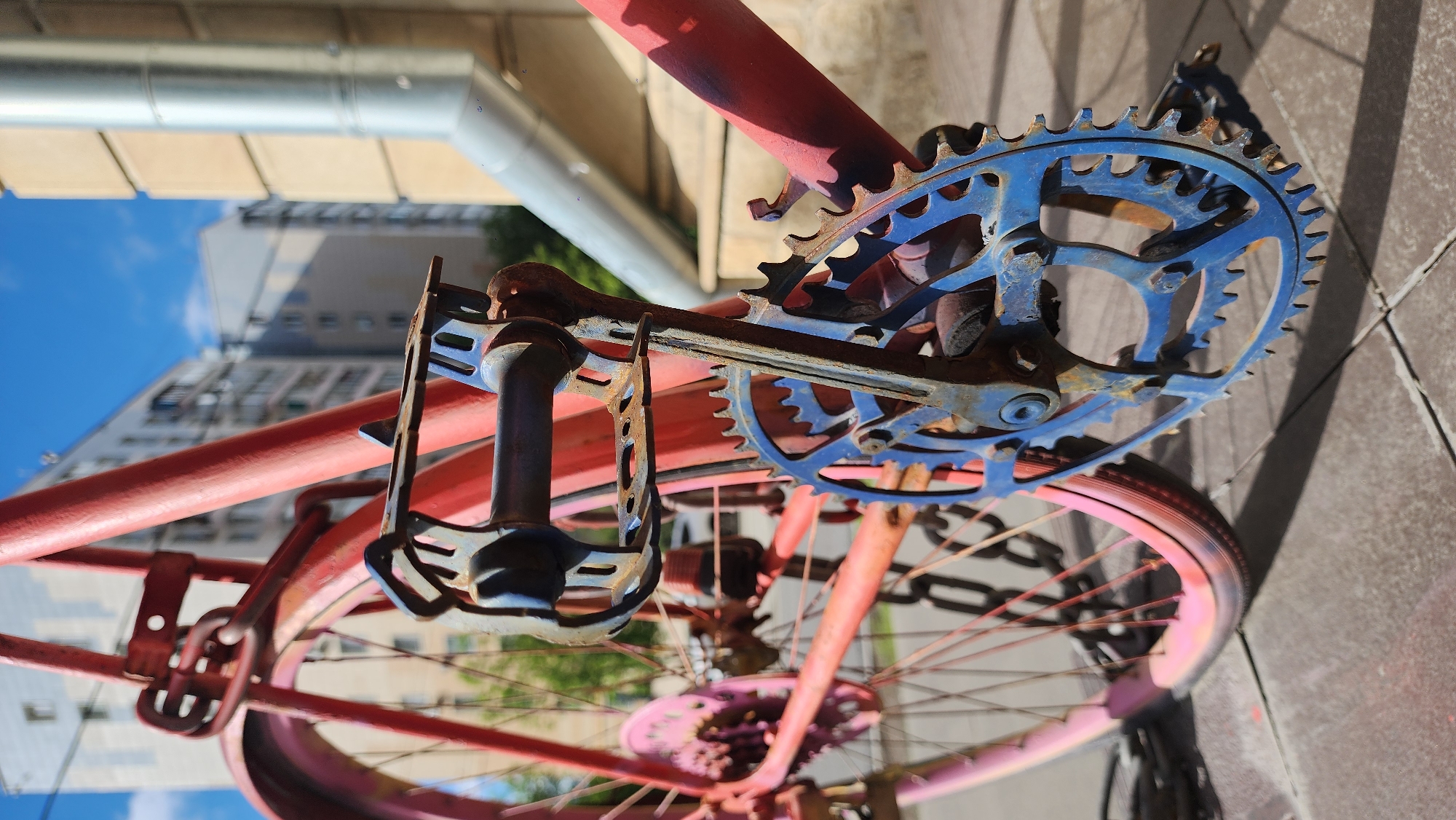 An upward-angled, eye-level shot depicts a portion of a red and pink bicycle, with a blue chainring and black pedal, positioned on a gray tiled surface. The image includes architectural elements such as a silver pipe above and a tall building visible through the bike frame, suggesting an urban setting. The scene is outdoors, illuminated by natural light, with shadows indicating the time of day. There are no people visible in the image. Given the information, it may be located in Moscow, Russia.