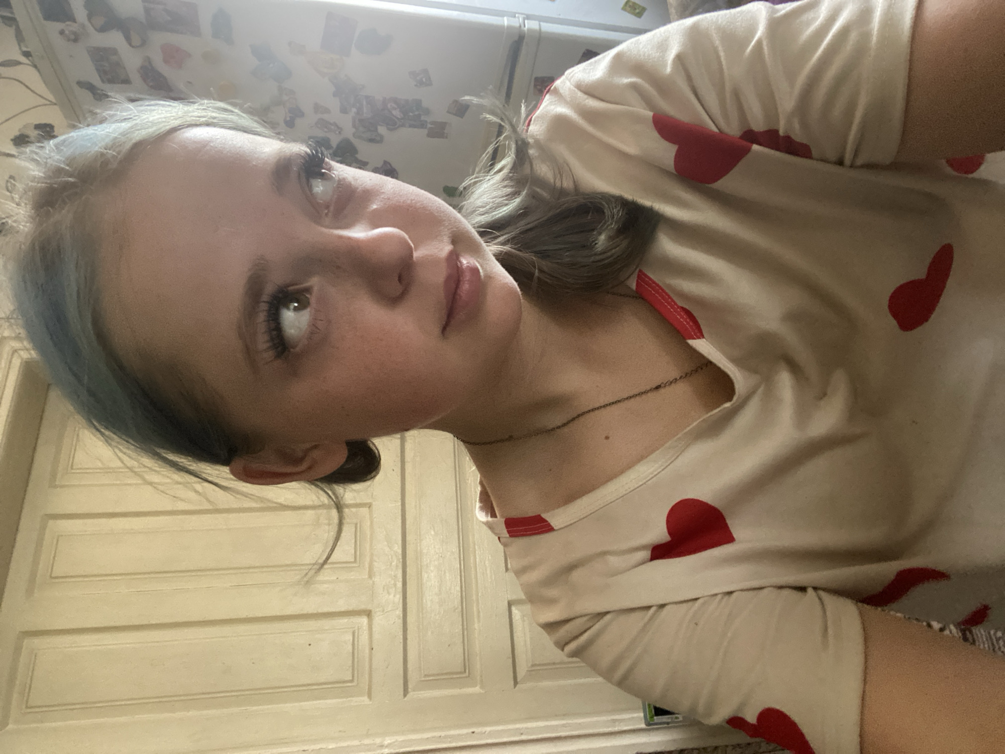 This is a close-up, over-the-shoulder, selfie-style photo of a young person, likely a girl, with light skin and blue-tinted hair. She is wearing a light-colored t-shirt with a repeating pattern of red hearts and a thin silver necklace. Her head is tilted, and she is looking upwards and to the left with a neutral expression. Her eyelashes are prominent, and she has a smattering of freckles across her nose and cheeks.
The background suggests an indoor setting. To the left, there are what appear to be framed pictures or items on a wall. To the right and behind her, there is a white door with paneling, indicating a room in a house or apartment. The lighting is bright, suggesting daytime, possibly with natural light entering the room.
The image is oriented with the top of the head on the left side of the frame. The overall impression is a casual, personal snapshot. There is no visible text in the image.
