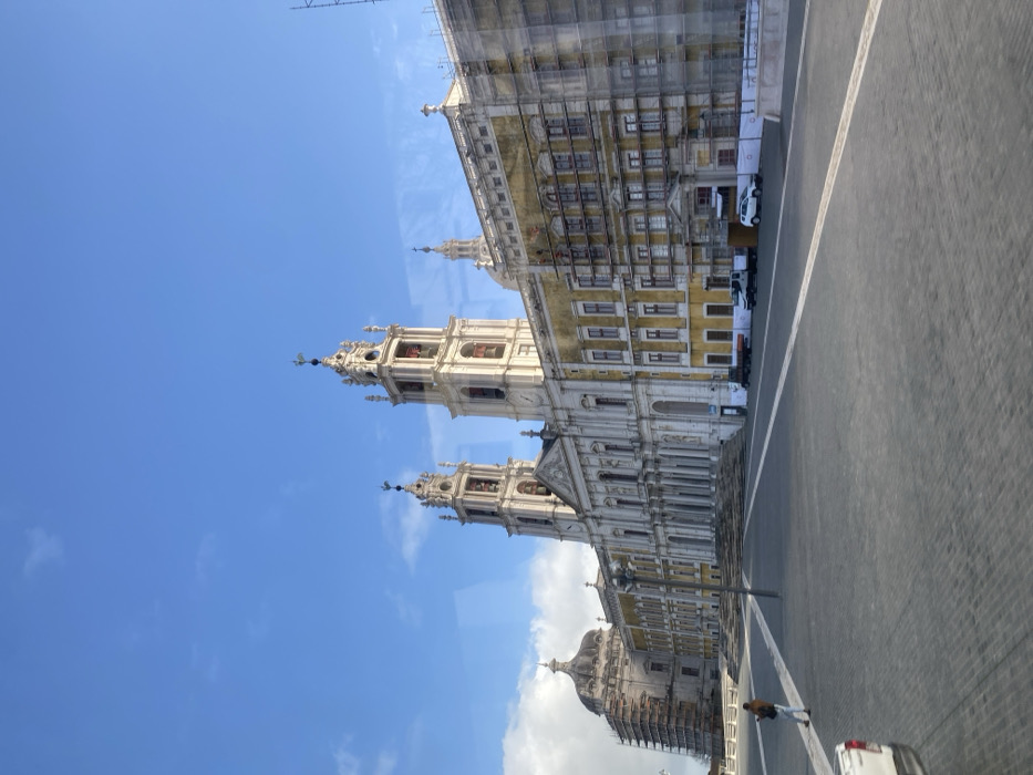The image captures a wide view of the Mafra National Palace in Mafra, Portugal, under a clear blue sky with sparse white clouds. The grand, Baroque-Neoclassical structure occupies the midground and background, featuring a prominent central basilica flanked by two tall, light-colored bell towers with decorative spires and visible bells. A large, paved plaza of dark grey pavers stretches across the foreground and midground.
The palace complex extends horizontally, with a yellowish facade on the right side and a lighter facade on the left. Significant portions of the building are undergoing renovation; the right wing and a dome structure on the far left are covered in extensive scaffolding. A wide set of stairs leads up to the main entrance of the central basilica.
In the plaza, a single individual is walking towards the left in the lower-left quadrant, wearing a brown jacket and light-colored pants. A white pickup truck is partially visible in the bottom-left corner. Near the base of the renovated right wing, several vehicles, including multiple trucks and a white car, are parked behind white barriers. A tall, dark pole, possibly a street light, stands in the midground of the plaza. Subtle reflections on the image surface suggest it may have been captured through a window.
