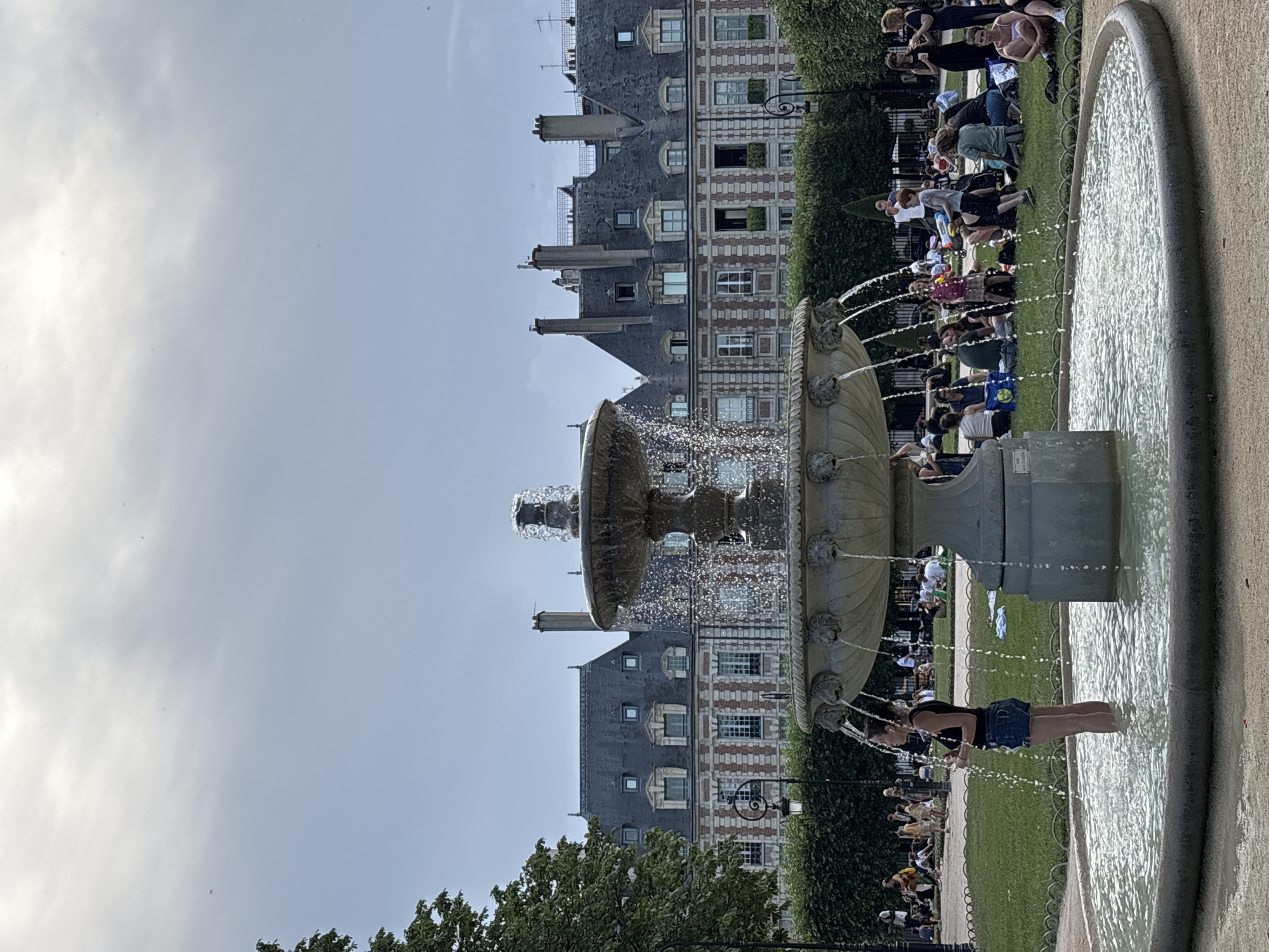 A woman stands with her legs submerged in the water of a large, multi-tiered stone fountain in the foreground. She is wearing a black tank top and denim shorts. The fountain is active, with water cascading from its upper levels. Behind the fountain, people are gathered on a grassy lawn, many sitting or lying down, appearing to be picnicking or relaxing. A row of ornate, multi-story buildings with dark tiled roofs and numerous windows lines the background. Green trees and hedges are visible on the left and right sides of the frame, framing the scene. The sky above is overcast with a bright, diffused light. This appears to be the Place des Vosges in Paris, France, a historic square with a central garden and fountain surrounded by arcades and classical architecture.