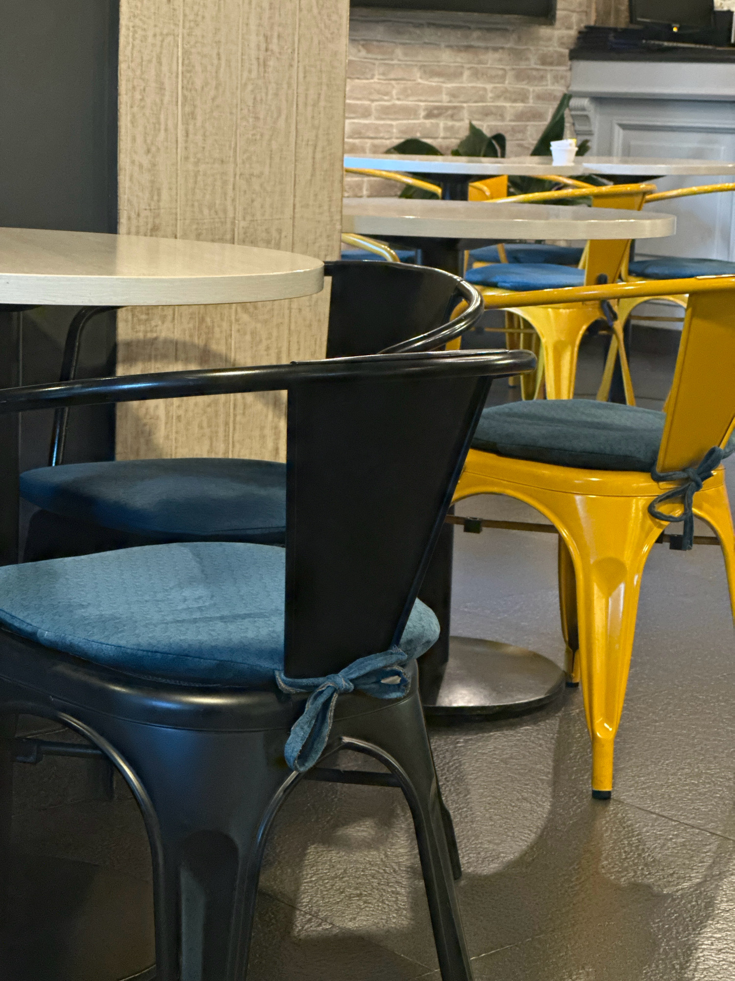 The image depicts a close-up of chairs and tables in what appears to be a cafe. In the foreground, a black chair with a blue cushion is prominently featured. To its right, a bright yellow chair with a similar blue cushion is visible, tied with a blue fabric strip. Further back, more yellow chairs and tables are discernible, with a hint of a brick wall in the background. The setting is indoors, with a textured floor and soft lighting, creating a cozy atmosphere. The arrangement of furniture suggests a dining or seating area. The image does not provide any specific clues that would allow for identification of the location within Kathmandu Metropolitan City, Nepal.