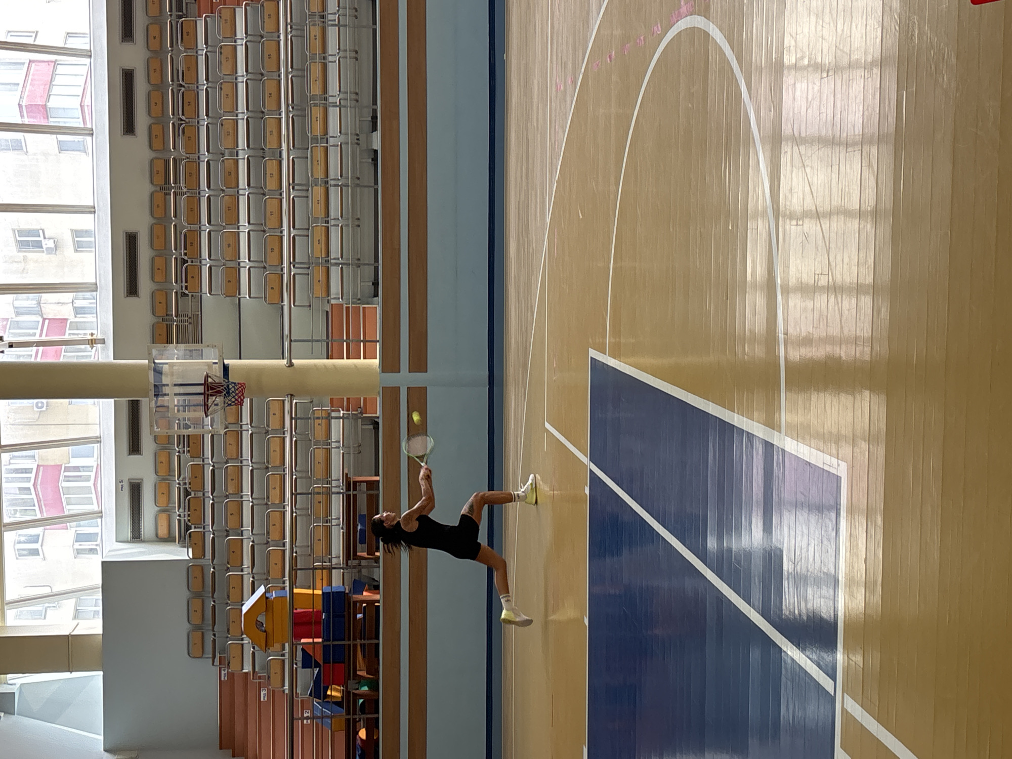 The scene is set inside a brightly lit indoor sports gymnasium in the city of Chaikovsky, Russia. A single individual, appearing female, with dark hair, is captured mid-action on a polished wooden court. She is wearing a black sleeveless top, black shorts, white ankle socks, and light-colored athletic shoes. A tattoo is visible on her left thigh. She is lunging forward to her right, holding a tennis racket with both hands, positioned to strike a yellow tennis ball suspended in mid-air directly in front of the racket head.
The gymnasium floor features dark blue painted sections on the left, delineated by white lines, suggesting a multi-sport court. Faint pink, curved markings are present on the lighter wooden section of the court to the right. In the background, tiered bleachers with numerous individual, light-colored seats (appearing yellow or orange) are visible, each with a metal frame and numbered seatbacks (e.g., "1" through "17"). On the lowest level of the bleachers on the far left, stacked colorful geometric shapes, resembling soft play equipment (red, blue, yellow), are present. A basketball hoop and backboard are partially visible on the left, behind a large circular pillar. The upper background features large, multi-paned windows extending across the entire width of the frame, through which multi-story residential buildings with various window patterns and some red-accented balconies are visible in daylight. Below the windows and behind the bleachers, a light blue wall section with a horizontal wooden trim runs across the gym. Rectangular ventilation grilles are visible high on the wall. The polished floor reflects the overhead lighting and the outdoor scene.