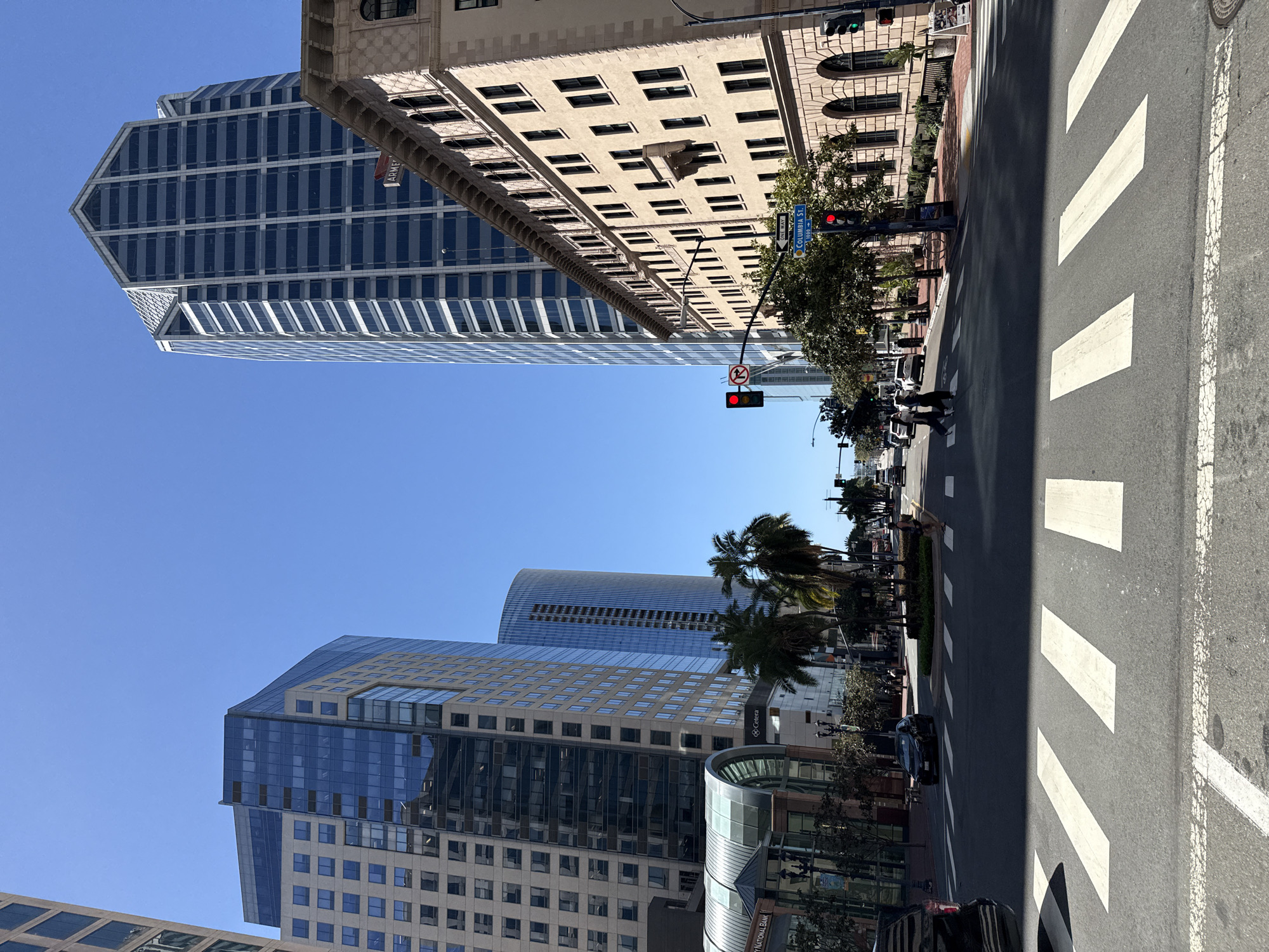 The image depicts a street scene in downtown San Diego, California. The view is looking east on B Street at the intersection of Columbia Street. Skyscrapers line the street, their glass and steel facades reflecting the clear blue sky. The Columbia Street sign is visible on a traffic light pole. Pedestrians are crossing the street in the crosswalk. A car is approaching the intersection. Palm trees add to the San Diego ambiance.