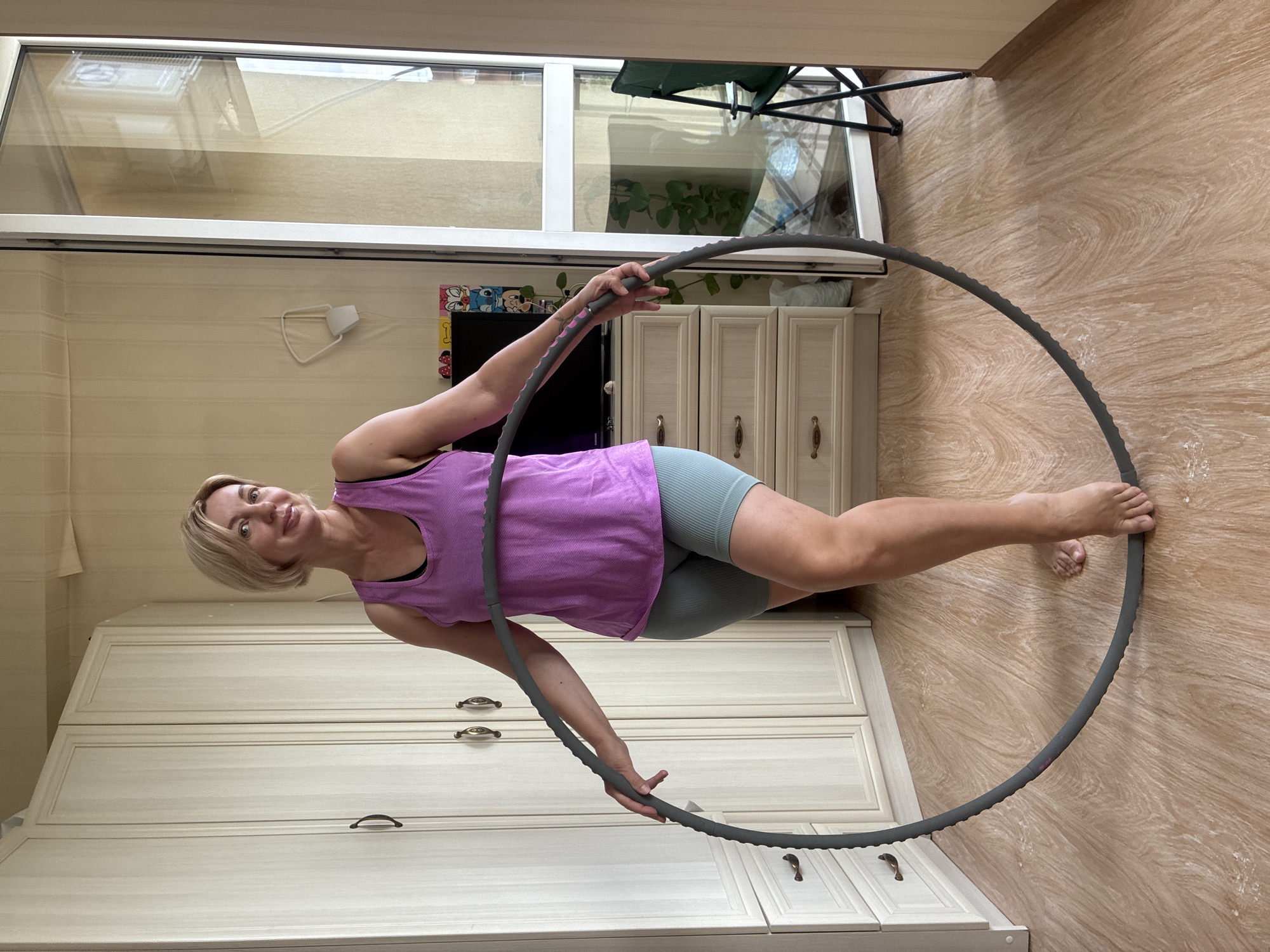 A blonde woman is standing indoors, holding a hula hoop. She is wearing a purple tank top and blue athletic shorts. The room has light-colored walls and wooden flooring. In the background, there is a large wardrobe, a dresser with drawers, and a television on top of the dresser. To the right, there is a glass door leading to a balcony, with a chair visible outside. The scene appears to be an apartment.