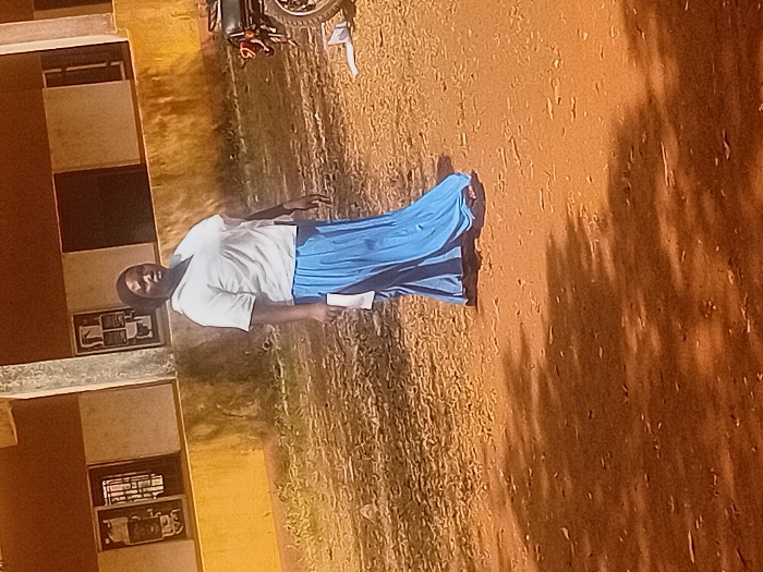 A woman stands in a schoolyard, dressed in a white shirt and long blue skirt. She holds a white paper. Nearby, a building with windows and mounted items is visible. The ground is a dry, reddish-brown color with sparse vegetation. Dark shadows are cast on the ground, possibly from trees or other structures. A motorcycle is parked near the top of the frame. The overall impression is of a rural or less developed setting.