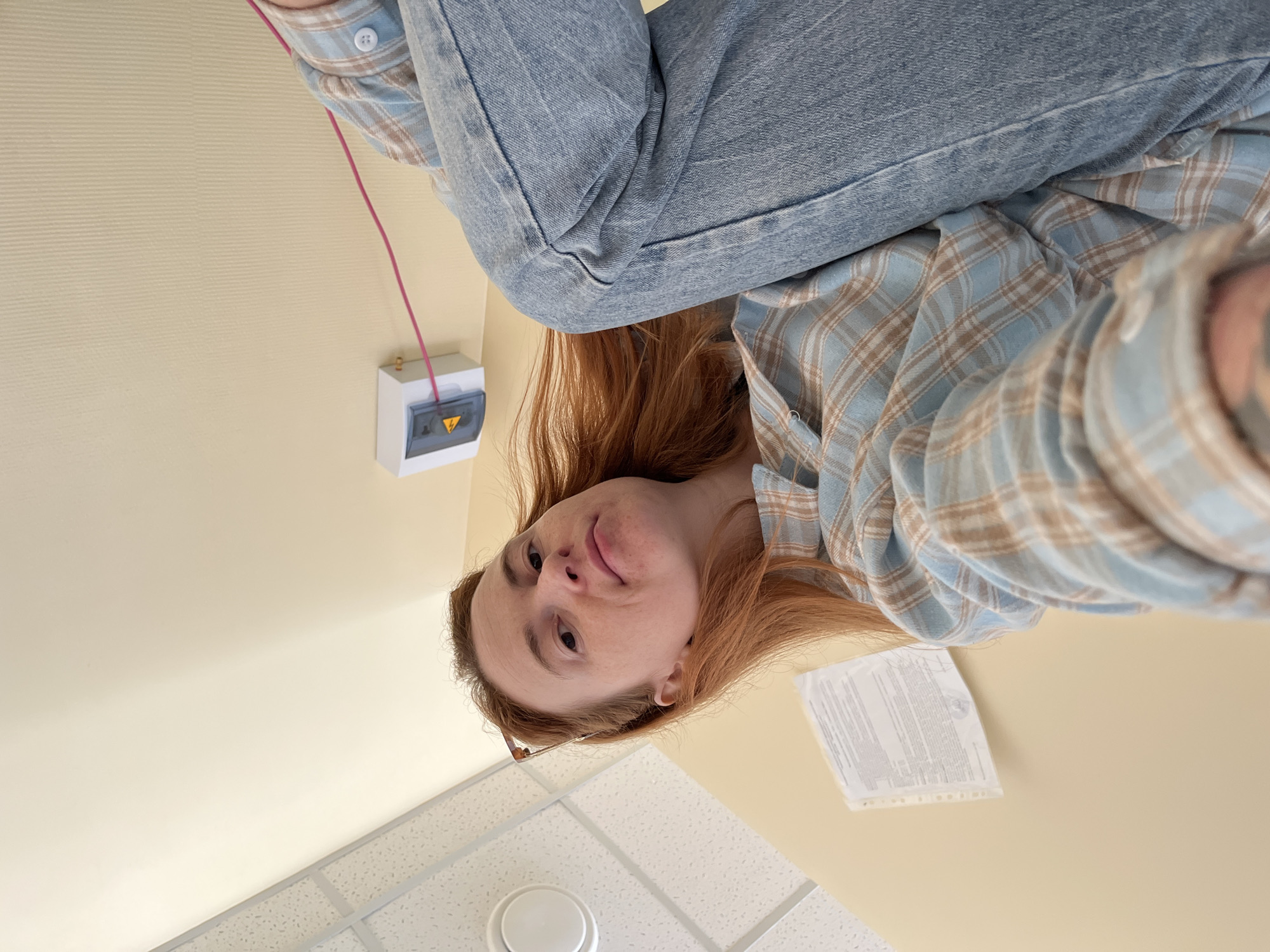 The rotated image, taken from a low, upward angle, features a young woman in an indoor setting. She has long, reddish-brown hair and looks towards the camera with a neutral to slightly inquisitive expression, showing a hint of a subtle smile. She is dressed in a light blue and brown plaid long-sleeved shirt and blue denim jeans. The frames of what appear to be glasses are partially visible near her right ear.
The background consists of a light beige wall and a white acoustic tile ceiling with a visible grid. On the wall, a small white electrical box with a transparent cover is mounted. A thin pink wire extends from this box upwards, out of the frame. The box has a yellow warning sticker featuring a black lightning bolt symbol, indicating an electrical hazard. A circular white fixture, likely a light or smoke detector, is visible on the ceiling. To the right of the woman's head, a piece of white paper with dense, illegible text is affixed to the wall.
The photograph appears to be a close-up, informal portrait or selfie, capturing the woman from an unusual perspective. The indoor environment suggests a functional space, such as an office or public building. No specific location cues are visible to identify it as Chaikovsky, Russia, beyond it being an indoor setting.