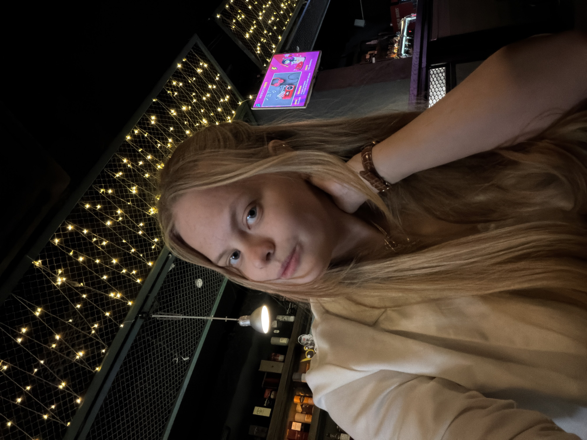 A woman with long blond hair is posing for a selfie indoors. She's wearing a light-colored sweater and a brown bracelet. Behind her is a wall covered in string lights. In the background, there are shelves of bottles, a monitor with colorful graphics, and other bar-like elements. The location appears to be a bar or restaurant, although the name of a city in Russia cannot be confirmed from the image.