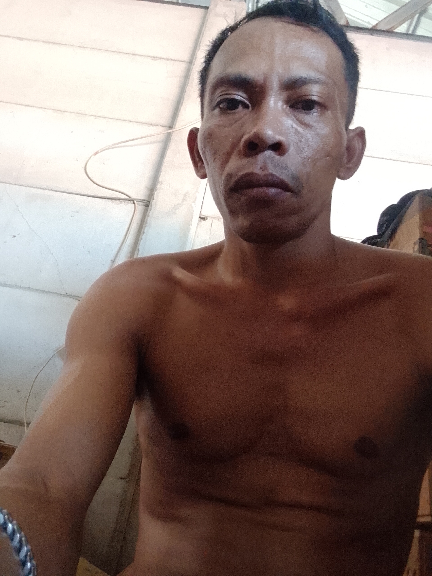 A shirtless man with dark hair and dark skin is centered in the frame. He appears to be sweating, and his expression is neutral. He is indoors, evidenced by a white ceiling and wall in the background. A wire is visible near the ceiling. The man is wearing a bracelet on his left wrist. The location is Bukit Ketok, Indonesia, according to the prompt. The scene is indoors, with focus on the man's upper body. The background is minimal and offers little additional context.