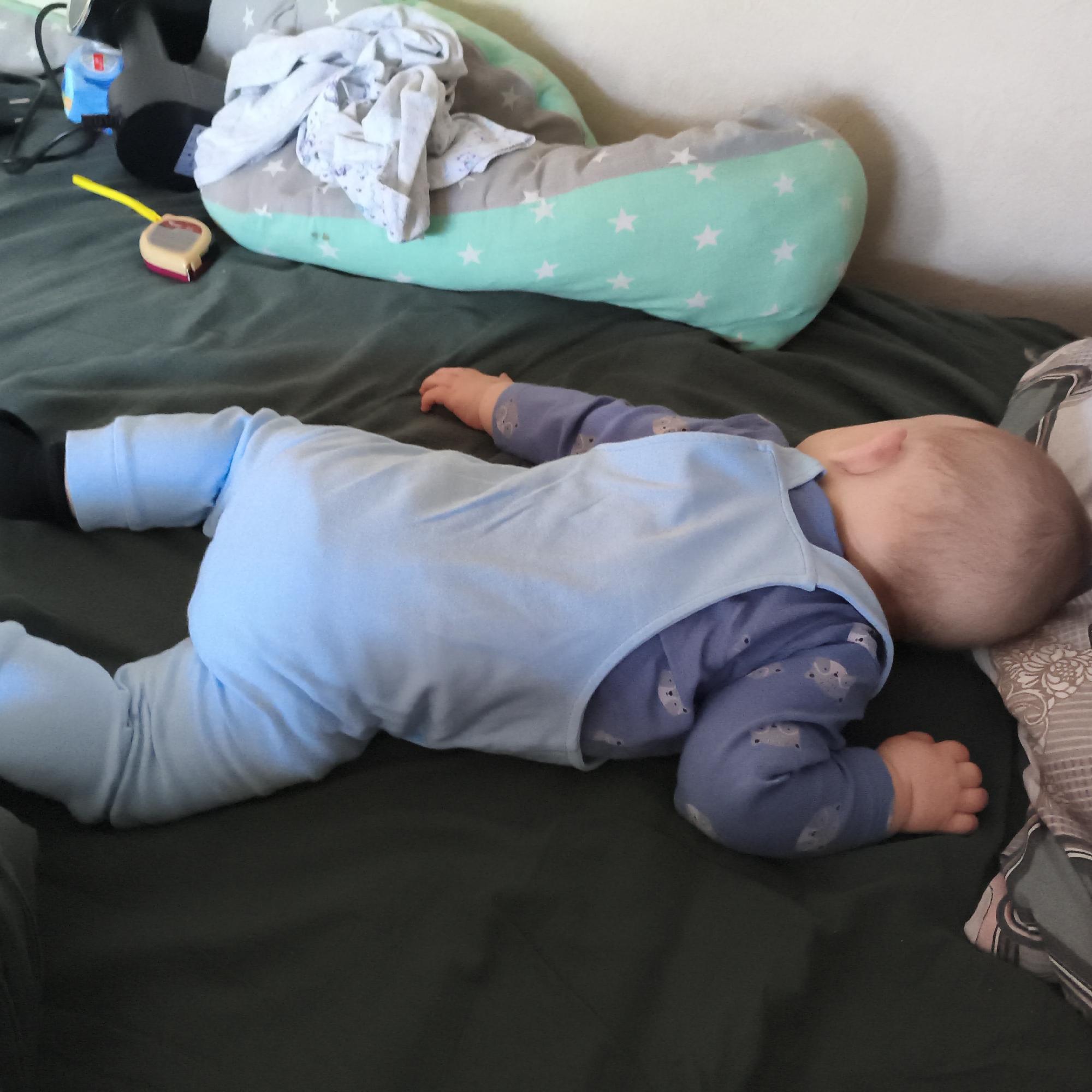 A baby is captured sleeping on its side on a dark green sheet or duvet cover. The baby is wearing a light blue sleeveless romper over a purple long-sleeved top patterned with small, light-colored animal figures (possibly elephants or hippos). Black socks are visible on the baby's feet. The baby's head is turned away from the camera, resting peacefully on the bedding.
In the background, to the left and behind the baby, is a large, crescent-shaped pillow. This pillow features a two-tone design, with sections of grey and mint green, both adorned with white star patterns. A pile of crumpled light-colored fabric, possibly baby clothes or a light blanket, is resting on top of this pillow.
Near the upper left of the image, a small toy resembling a yellow and red measuring tape lies on the dark green surface. Other blurred objects, including a dark shape and a blue item, are partially visible in the background, suggesting a lived-in indoor environment.
The scene depicts a calm and peaceful moment, likely indoors in a bedroom or living area in Chaikovsky, Russia. There is no visible text in the image.