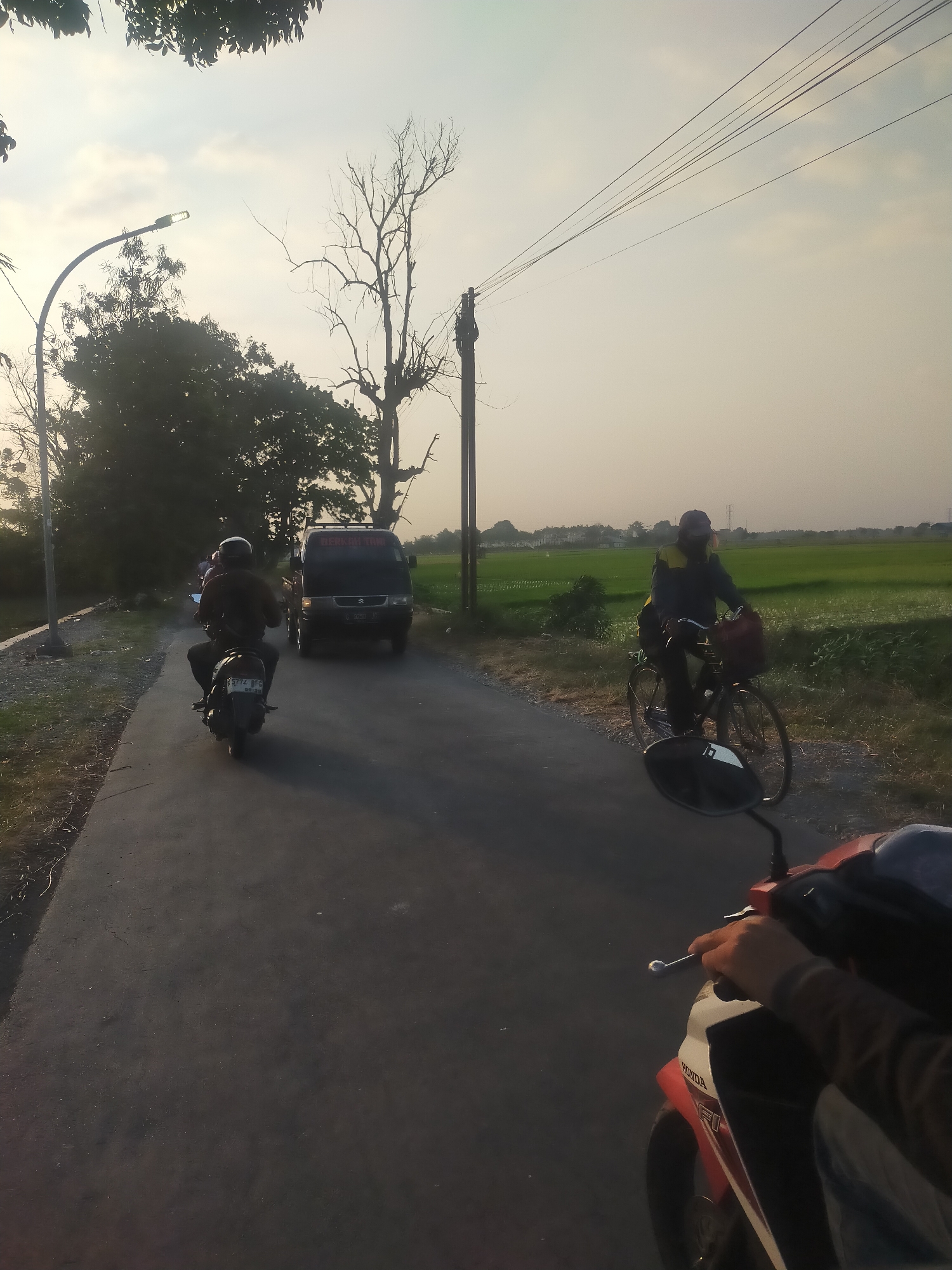 The image depicts a road scene in Dukuhdamu, Indonesia, at dusk. A dark asphalt road runs through a rural landscape with fields of green crops on the right and trees on the left. A motorcycle, ridden by a person wearing a dark helmet and jacket, is in the foreground, moving away from the camera. Further down the road, a dark van is traveling in the same direction, followed by another motorcycle. On the right side of the road, a person wearing a cap and a jacket is riding a bicycle, also moving away from the viewer. Power lines stretch across the sky, supported by utility poles. The sky is overcast with a hazy glow, suggesting the setting sun.