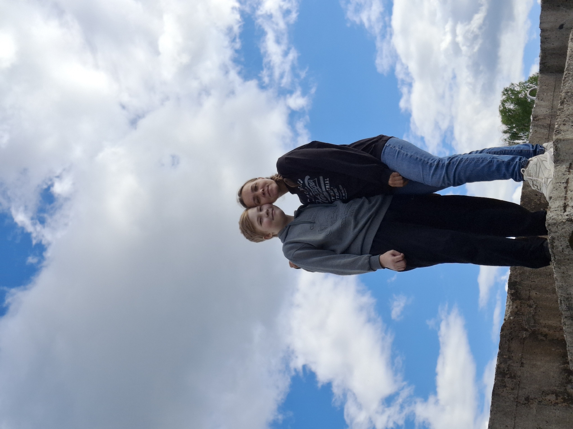 This media file features two young people posing for a photo against a bright blue sky with scattered white clouds. The perspective is from below, looking up.
The subject in the foreground, a younger person with blonde hair, is wearing a grey hooded sweatshirt and black pants. They are looking up and smiling. Resting their head on the shoulder of the foreground subject is another person, presumably older, wearing a black hooded sweatshirt and blue jeans. This person is also looking towards the camera and smiling. Their legs are extended upwards, resting on a concrete structure that runs diagonally across the right side of the frame. The concrete appears weathered and rough, suggesting an outdoor, possibly urban or industrial, setting. A glimpse of green foliage and a tree is visible in the distance, behind the concrete structure.
The lighting suggests it is daytime, likely mid-morning or afternoon, with bright sunlight illuminating the scene. The prominent blue sky and white clouds indicate fair weather. The overall mood is lighthearted and playful.
There is visible text on the black sweatshirt of the person in the background. While not entirely clear, some words appear to be present, possibly related to a brand or slogan.
The location context provided is Chaikovsky, Russia.
