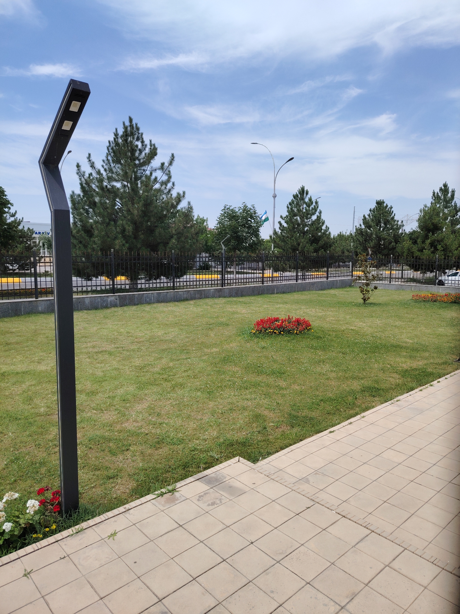 The image depicts a well-maintained park area with a paved walkway in the foreground. A dark, modern lamppost stands on the left side of the frame. The park features a lush green lawn with a circular flower bed containing red and yellow flowers. In the background, a black metal fence encloses the park, behind which are several tall pine trees. A street with yellow markings is visible beyond the fence, along with some buildings and parked vehicles. The sky is blue with scattered white clouds. The location is identified as Zaamin, Uzbekistan, as indicated by the Uzbek flag flying on a flagpole in the distance.