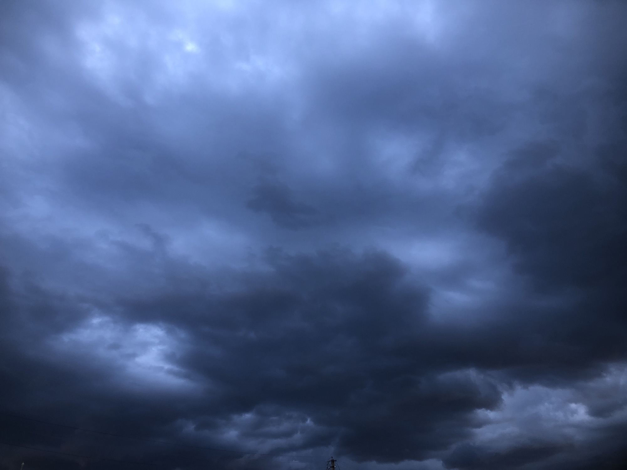 A wide shot shows a dark, stormy sky filled with heavy, ominous clouds. The clouds are a deep blue-grey, with lighter patches suggesting breaks in the dense formation. The lighting is dim, indicating it's either dusk or heavily overcast. The lower portion of the frame reveals a sliver of a dark horizon, with a faint hint of a pole or antenna structure visible at the very bottom center. No people or specific landmarks are discernible in this atmospheric view.