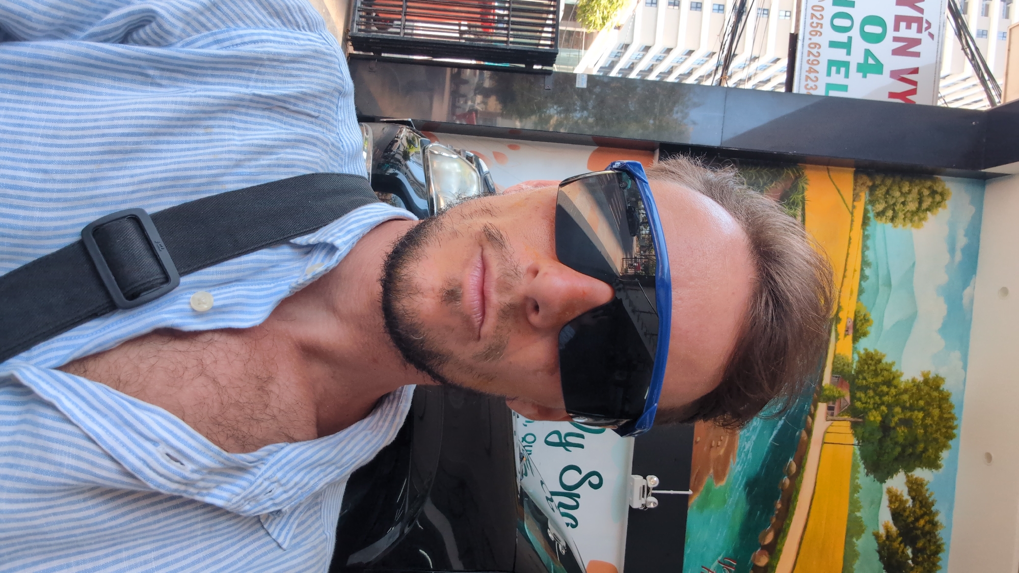 An adult male with fair skin, brown hair, stubble beard, and mustache is depicted in a close-up selfie in South Quy Nhon Ward, Vietnam. He wears blue-framed, dark-lensed sunglasses and a light blue and white striped button-up shirt, unbuttoned to reveal chest hair. A dark grey shoulder strap with a black buckle crosses his right shoulder. He faces the camera with a neutral expression.
The background on the left reveals a white sign with "YÊN VY 04 HOTEL" in red and green text, along with a partial phone number "0256.629423". Beyond the sign, a tall, light-colored urban building and a black metal gate are visible. To the right, a large mural on a wall depicts a rural scene featuring yellow fields, green trees, a winding blue river, a small house, and distant blue mountains. A dark-colored vehicle, likely an SUV, is partially visible behind the subject's right shoulder, showing its headlight and part of the grille. White text, "y Spa," is partially visible on the wall above the vehicle. The reflections in the subject's sunglasses show an urban street scene with vehicles and buildings.