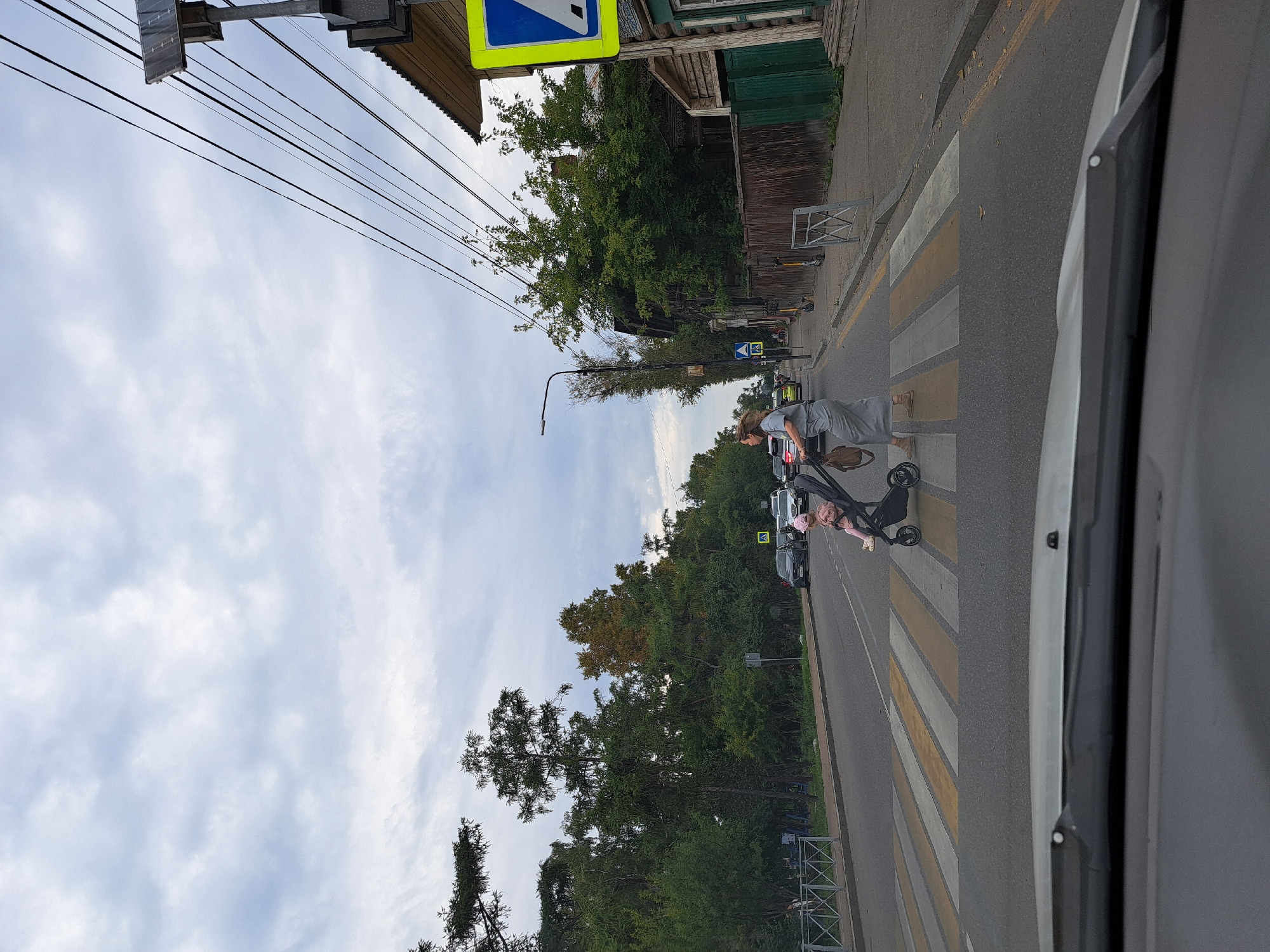 The image captures a street scene, likely in Irkutsk, Russia, under an overcast sky. A pedestrian crossing, marked with yellow and white stripes, is prominent in the foreground. A woman in a long grey dress is pushing a stroller with a child, dressed in a pink outfit and hat, across the street. Several cars are visible on the road, some stopped, suggesting adherence to pedestrian right-of-way. On the right side of the street, a wooden fence and buildings, one with a corrugated metal roof, are present. Overhead, power lines crisscross the sky. To the right of the road, a blue and yellow traffic sign indicating a pedestrian crossing is mounted on a pole. The scene appears to be on a residential street with trees lining the sidewalks and bordering the road. The perspective is from within a vehicle, looking forward through the windshield, with the lower edge of the windshield wiper visible.