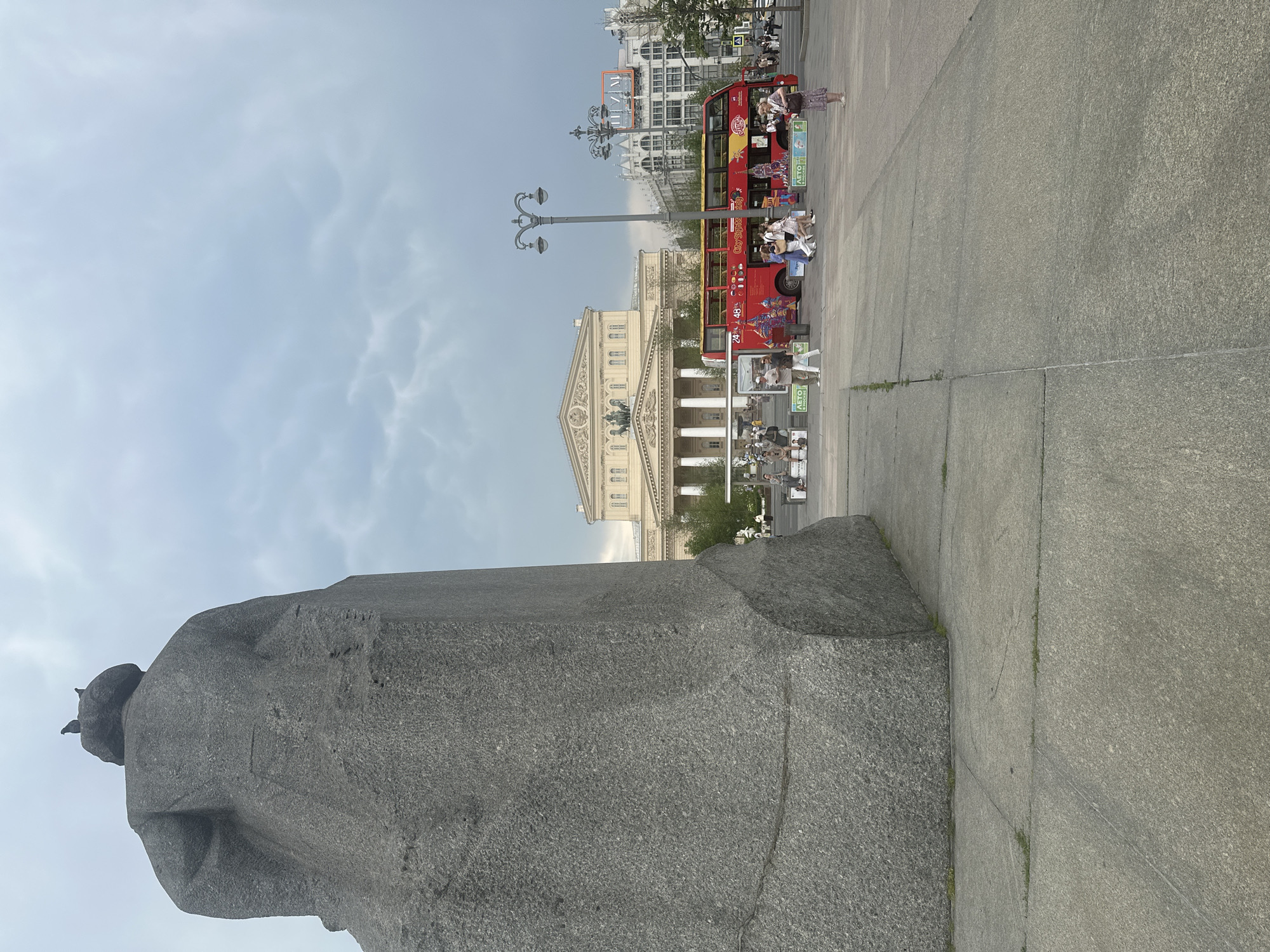 A large, rough-hewn granite monument occupies the left foreground of the image, obscuring much of the background. Two pigeons are perched atop the monument. In the middle ground, a red double-decker tourist bus with "City Sightseeing" branding is parked. Several people are walking around the bus and on the paved plaza in front of it. Behind the bus, the facade of a large, ornate building with classical columns and a pediment is visible, identified as the Bolshoi Theatre in Moscow, Russia. To the right of the Bolshoi Theatre, a multi-story building with intricate architectural details and windows can be seen. The sky above is overcast with heavy, grey clouds. The overall scene depicts a public square in Moscow on a cloudy day.