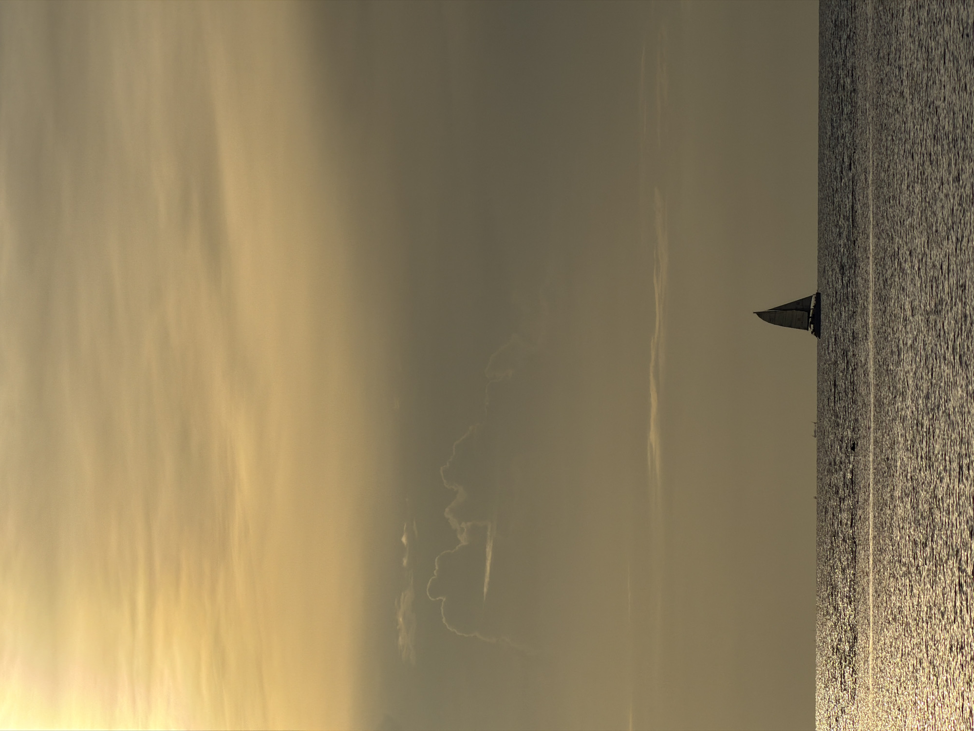 This media file depicts a serene seascape at sunset. The scene is dominated by the vast expanse of the ocean, with sunlight reflecting off its rippled surface, creating a textured, shimmering effect. The sky above is a hazy blend of warm, golden and muted grey tones, suggesting a dramatic sunset with scattered, wispy clouds.
A solitary sailboat is visible in the middle ground, its dark silhouette a stark contrast against the bright water and sky. The sailboat appears to be sailing gently, its sail catching the light. The overall mood is peaceful and tranquil, conveying a sense of calm and solitude. The lighting indicates it is late afternoon or early evening, with the low angle of the sun casting long shadows and bathing the scene in a soft, warm glow. The location cues, such as the tropical appearance of the water and sky, align with the provided context of Baan Bo Phut, Thailand. There is no visible text in the image.