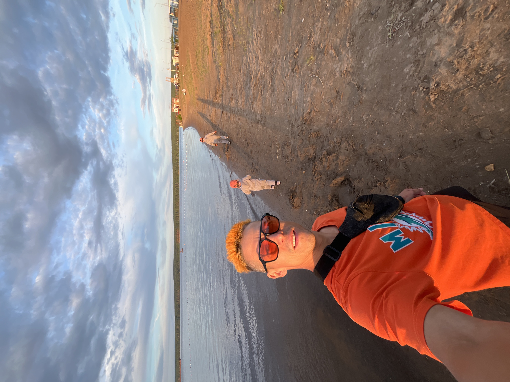 The image shows a person with bright orange hair and sunglasses taking a selfie outdoors. They are wearing an orange t-shirt with a logo on it that appears to say "MIAMI" with a dolphin design. A dark bag is slung across their chest. The setting is a beach or shoreline with calm water to the left and muddy sand to the right. In the background, there are a few other people dressed in light-colored outfits, appearing to be walking along the shoreline. There are also some structures visible in the distance on the land. The sky is filled with dramatic clouds, suggesting it might be late afternoon or early evening, and the lighting indicates a setting sun. The overall mood seems relaxed and casual.