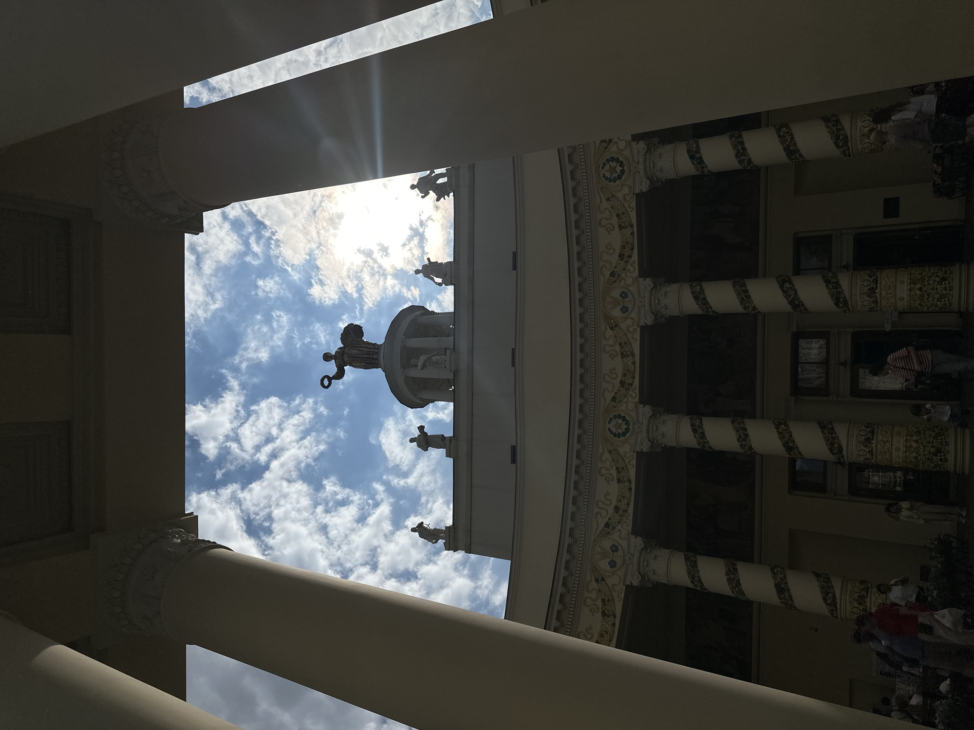 The image shows a building with classical architecture in an unknown city. The building has large columns and a statue on the roof. The sky is visible through the openings in the building. People are walking around outside the building.