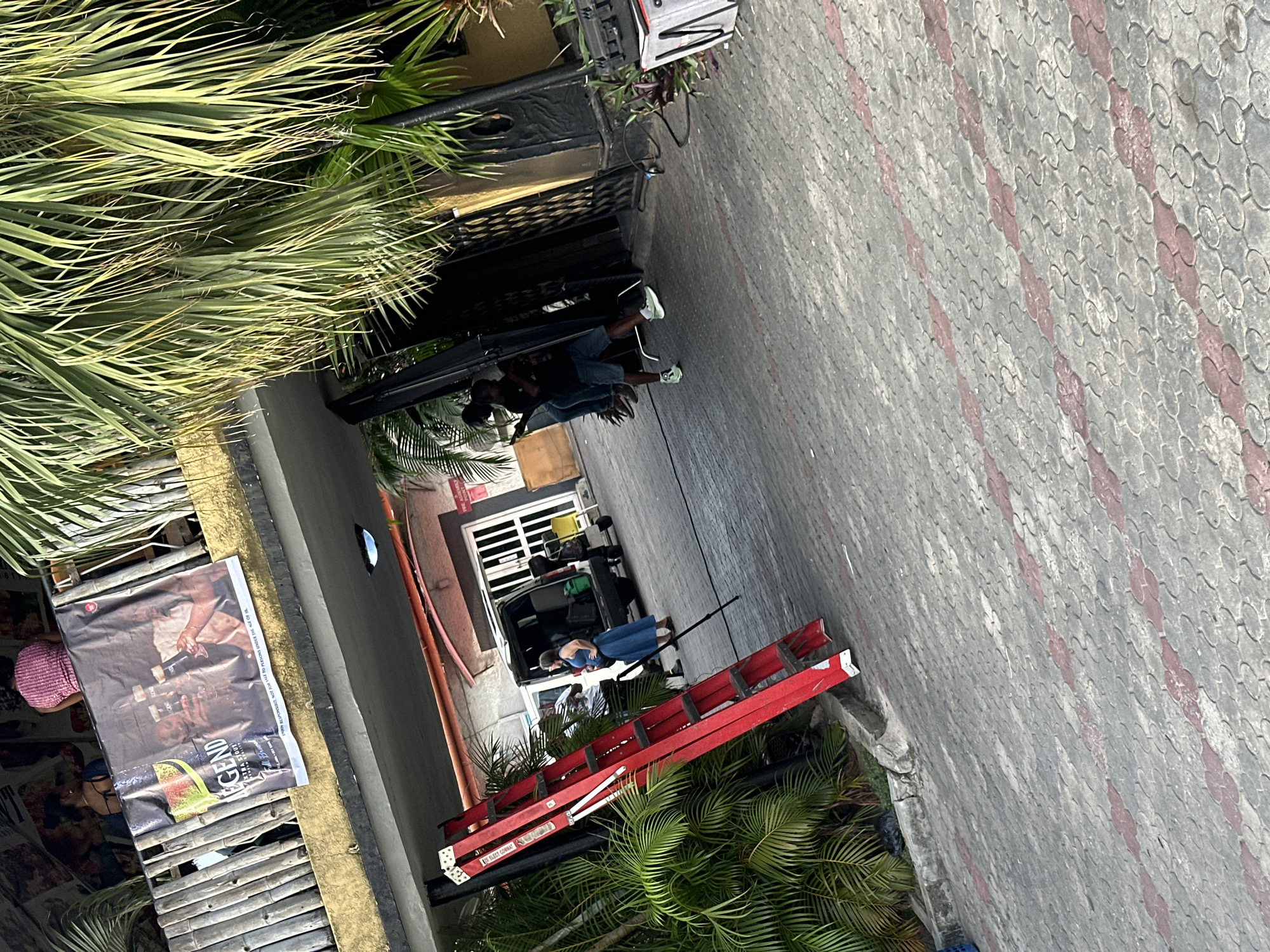 The image shows an outdoor scene in Lagos, Nigeria, featuring a red ladder leaning against a building. A woman in a blue dress is standing near the ladder. A group of people is near a van, appearing to be loading equipment. A banner with the word "LEGEND" is affixed to a bamboo structure. Palm trees are visible in the foreground. The ground is paved with grey and red bricks.