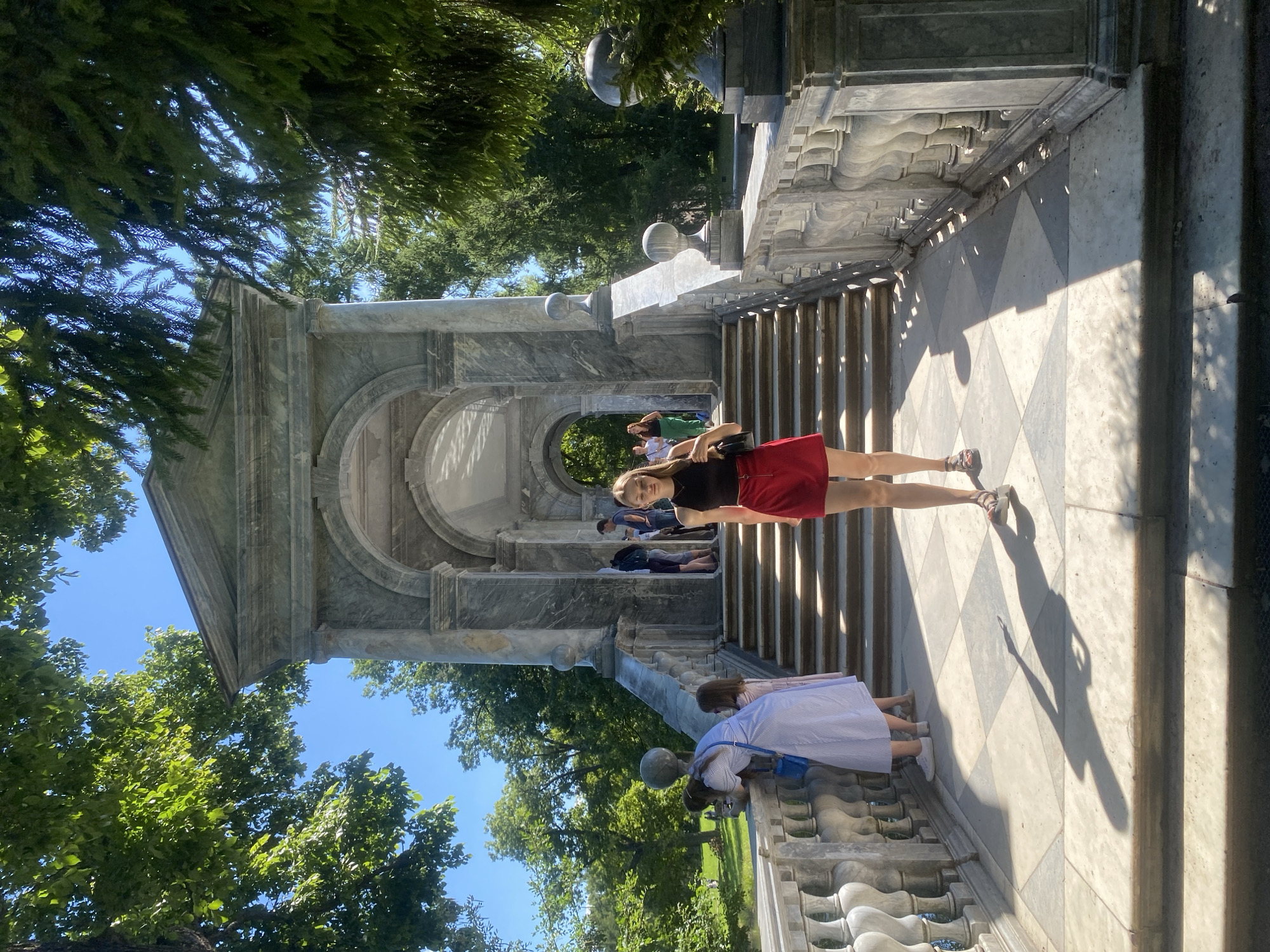 A young woman stands on a checkered paved area in front of a marble structure with arched openings and a grand staircase. She is wearing a black sleeveless top and a red skirt, holding a black purse. Sunlight casts shadows on the ground. In the background, several other people are visible near the stairs and in the distance, some of whom are leaning on a marble balustrade. The environment appears to be a park with lush green trees and a clear blue sky, suggesting a warm day. The marble structure is likely part of a palace or park ensemble in Pushkin, Russia, possibly Catherine Park given the architectural style.